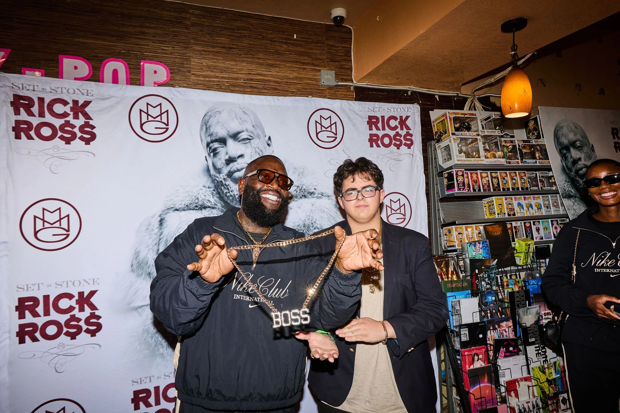A group of people, including Rick Ross, posing at an event with a banner behind them that has Rick Ross's name and logo. They are surrounded by shelves of Funko Pop figures and other merchandise.