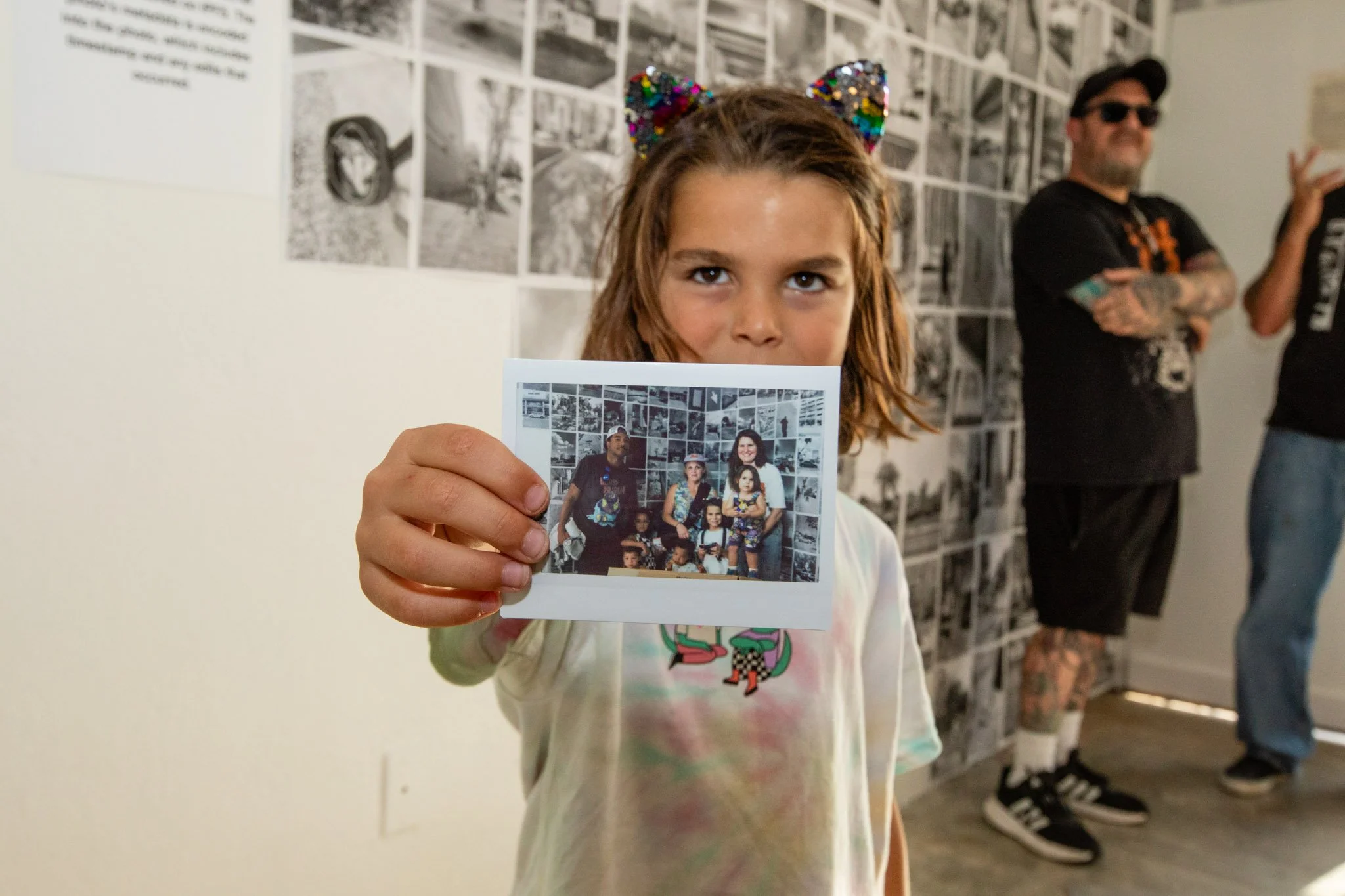 Young girl with cat ear shaped headband holding a photograph in front, showing a family photo in an art gallery with black and white pictures on the wall. Two men with tattoos and dark clothing are in the background.