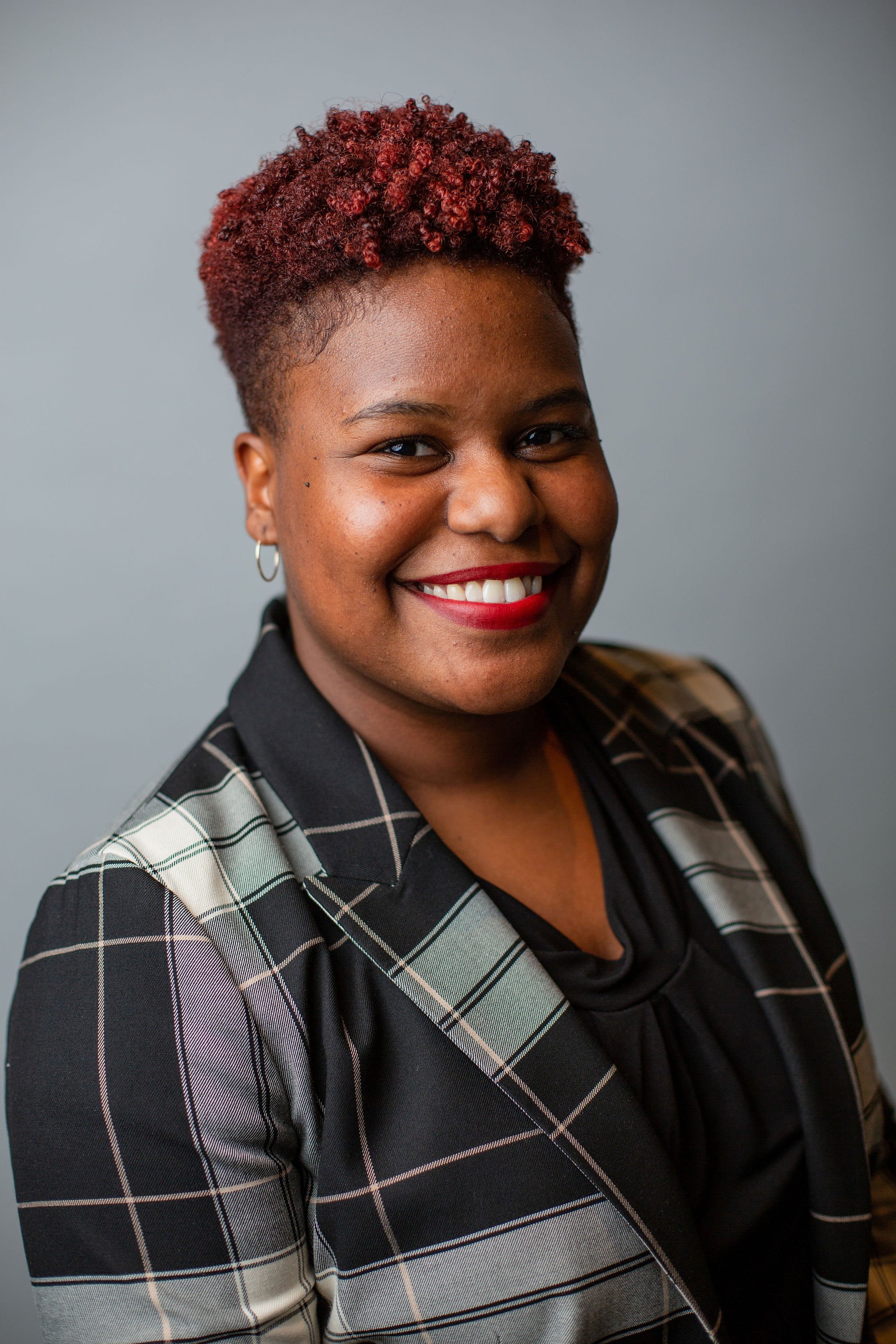 A woman with dark skin and curly red hair, wearing a black top and a black and white plaid blazer, smiling against a neutral background.