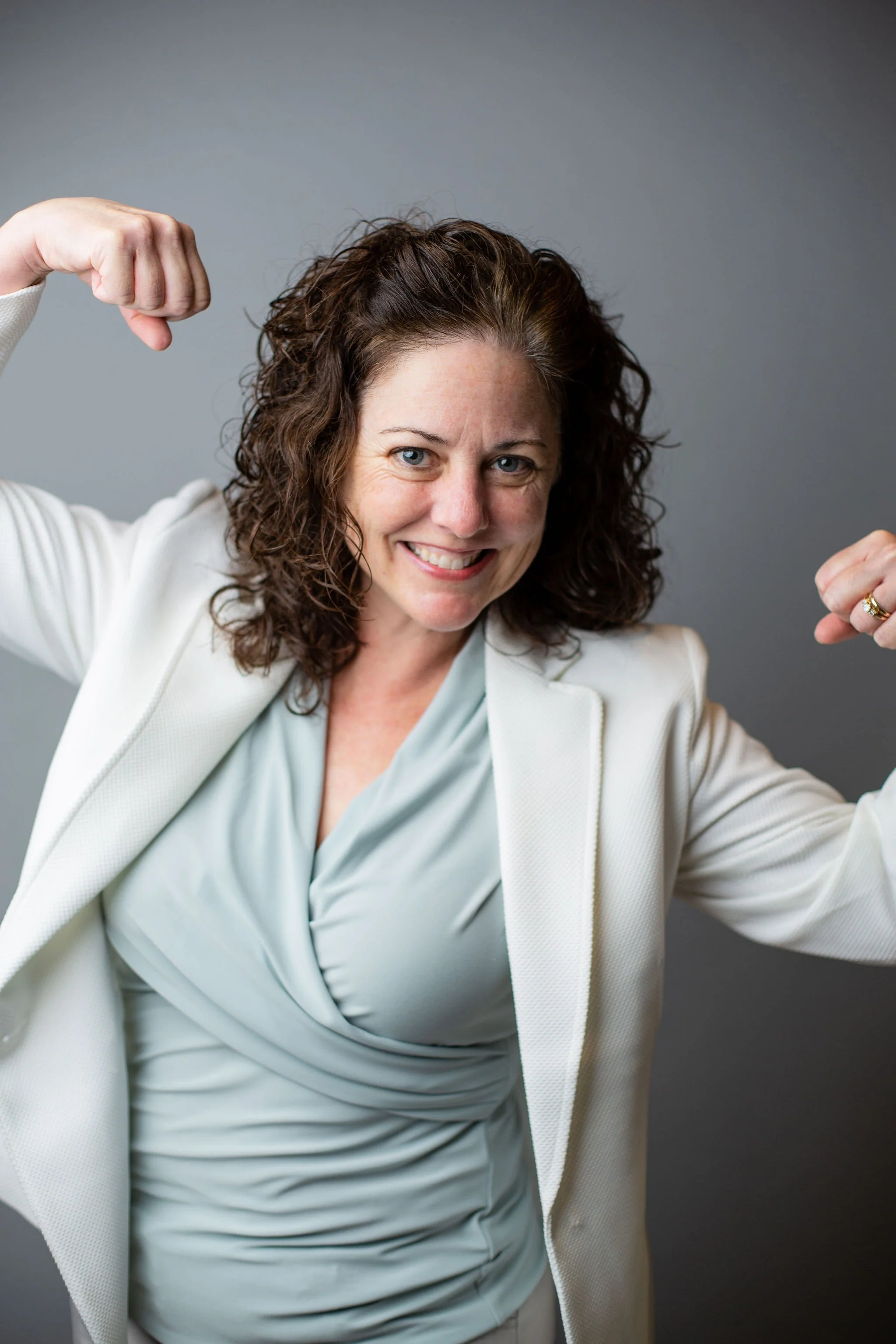 A woman with curly brown hair smiling and flexing her biceps, dressed in a light-colored blazer and blouse, standing against a plain gray background.