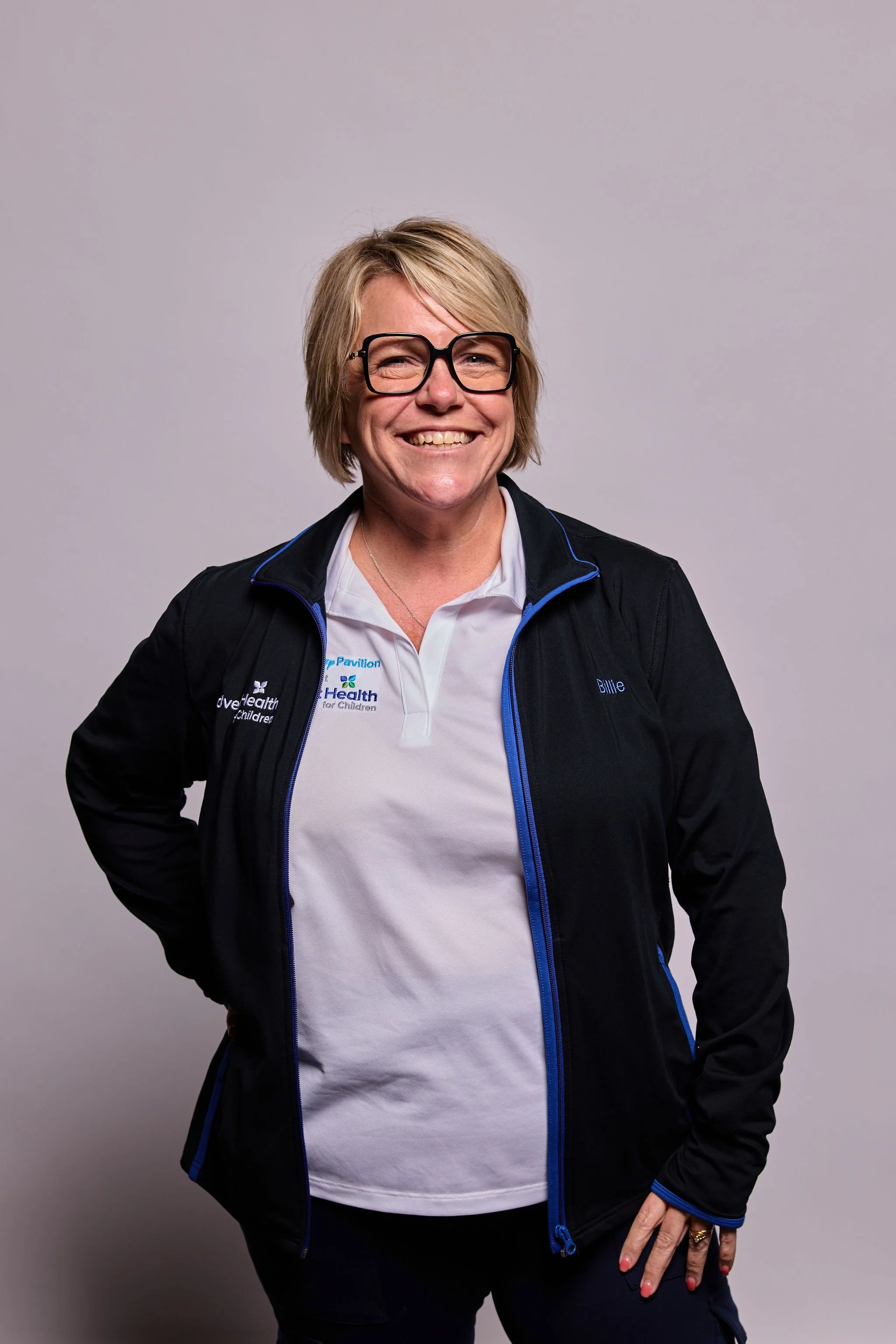A middle-aged woman with short blond hair wearing glasses, a black jacket with blue accents, and a white polo shirt with logos. She is smiling, standing with one hand on her hip against a plain gray backdrop.