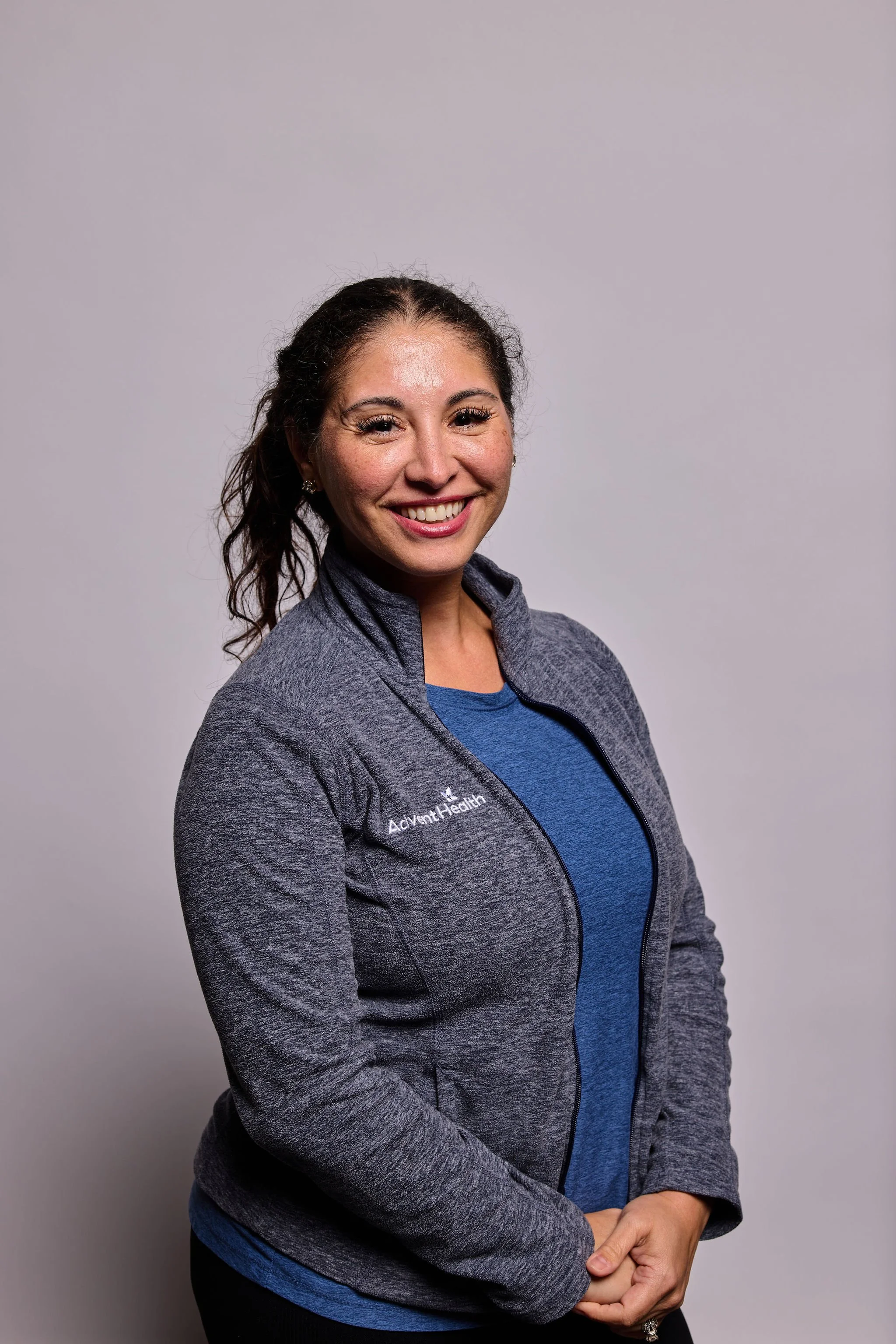 A woman smiling with her arms crossed wearing a gray jacket and blue shirt against a plain background.