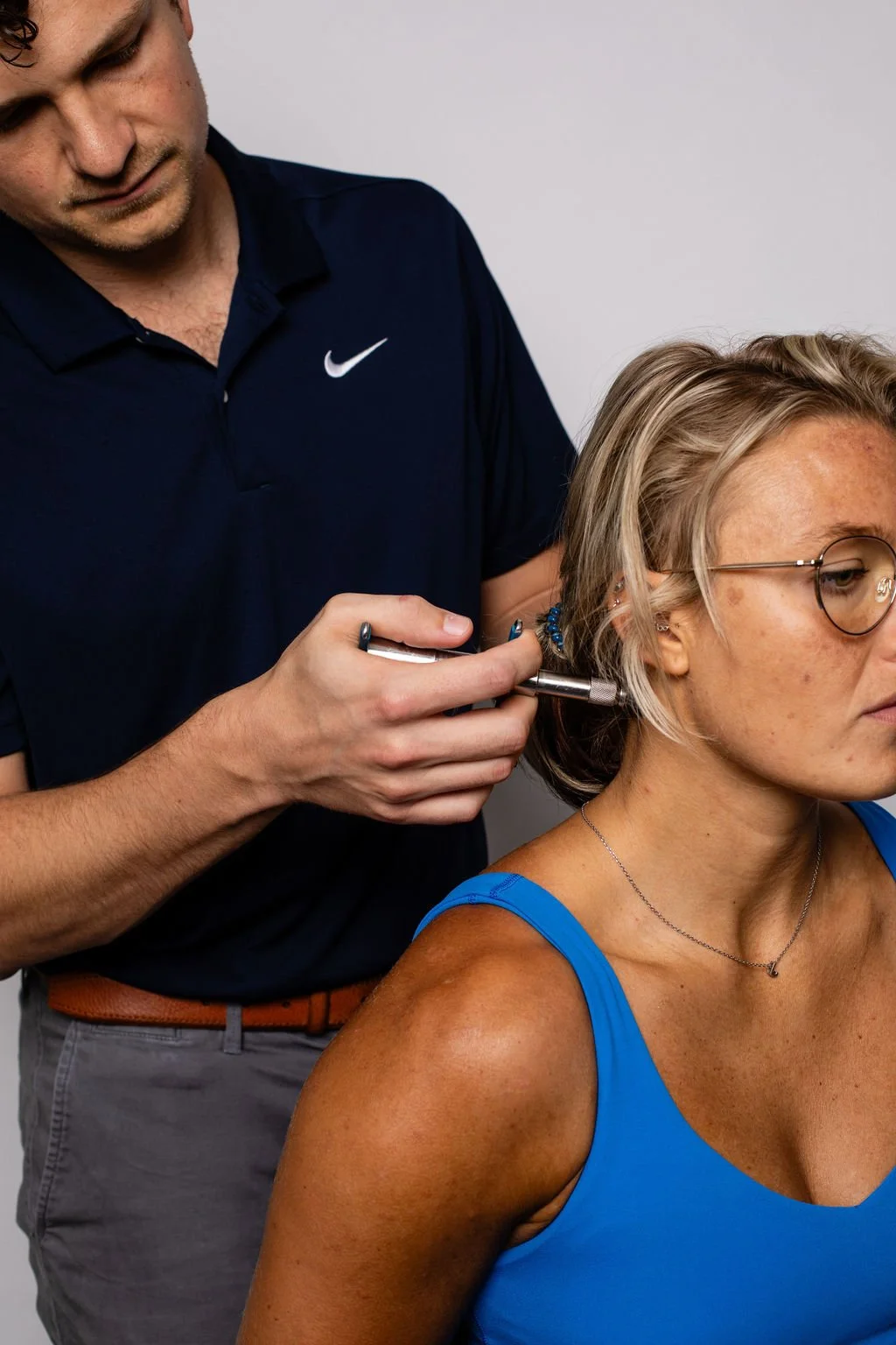A man in a navy Nike polo shirt and gray pants is holding a tool near a woman's ear, who is wearing glasses, a blue tank top, and a necklace, while seated against a plain gray background.