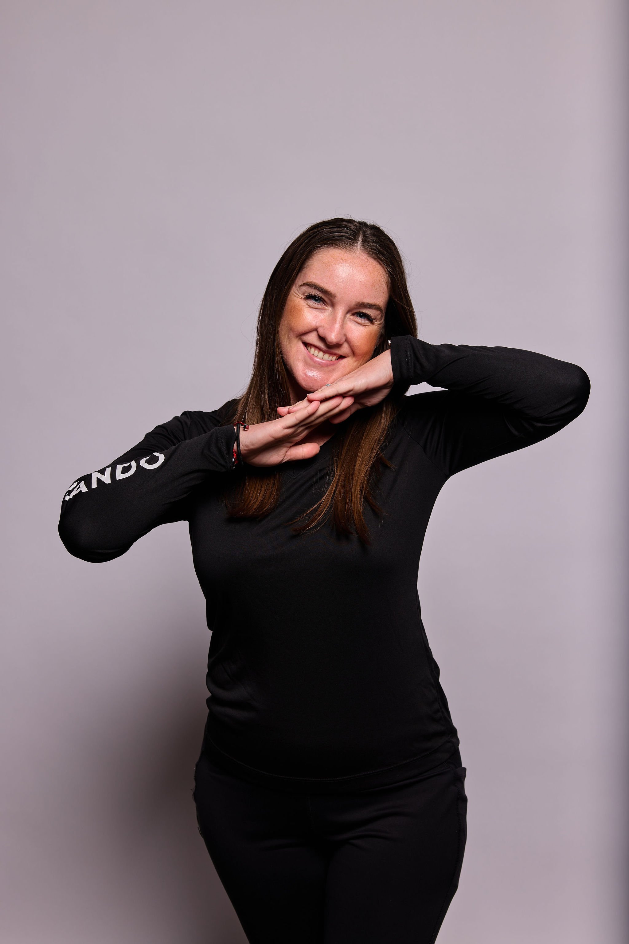Young woman with long brown hair wearing a black long-sleeve shirt and black pants, smiling and resting her chin on her folded hands in front of a plain light gray background.