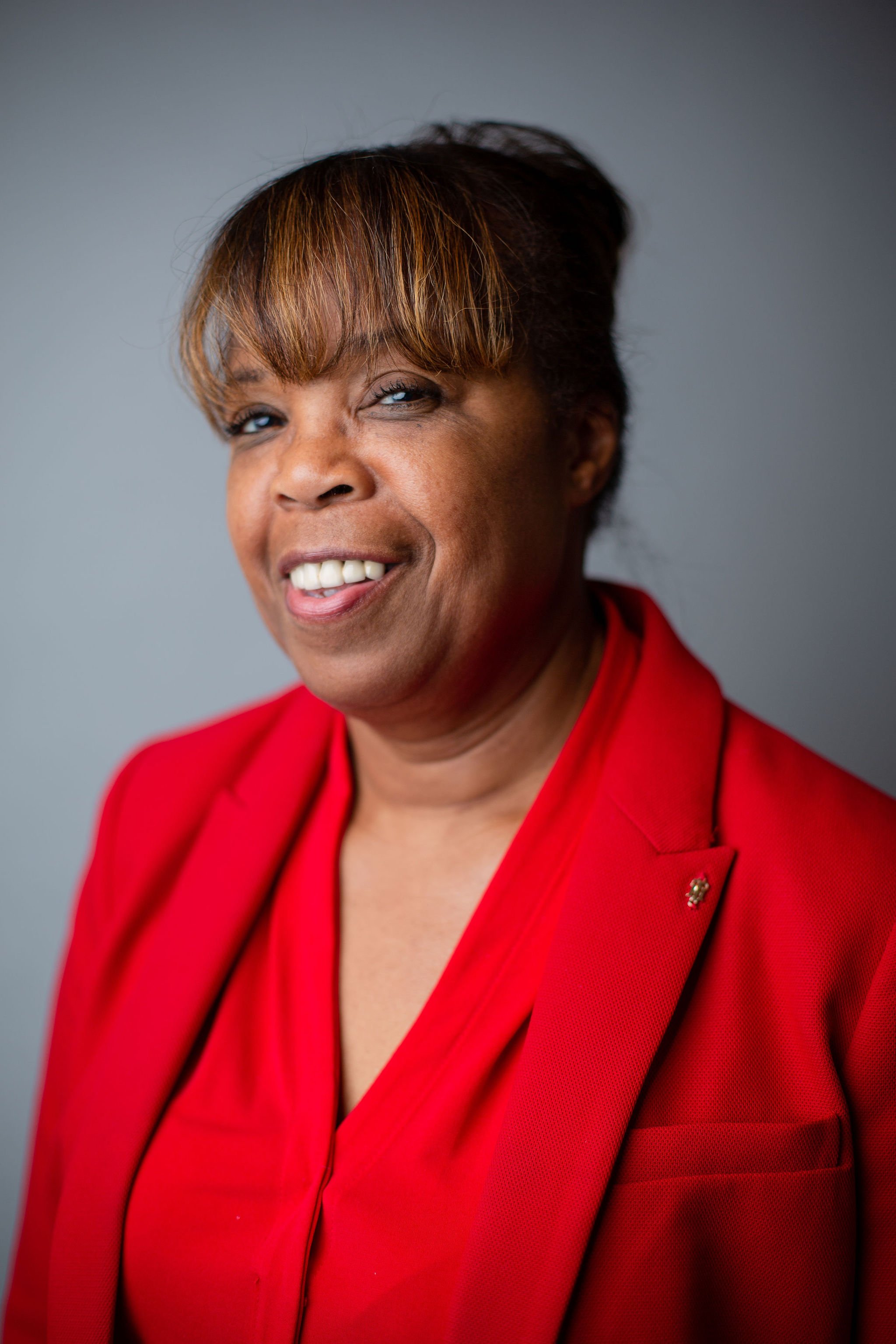 A middle-aged African American woman with short brown hair and bangs, smiling, wearing a red blazer with a small pin on the lapel, against a neutral grey background.