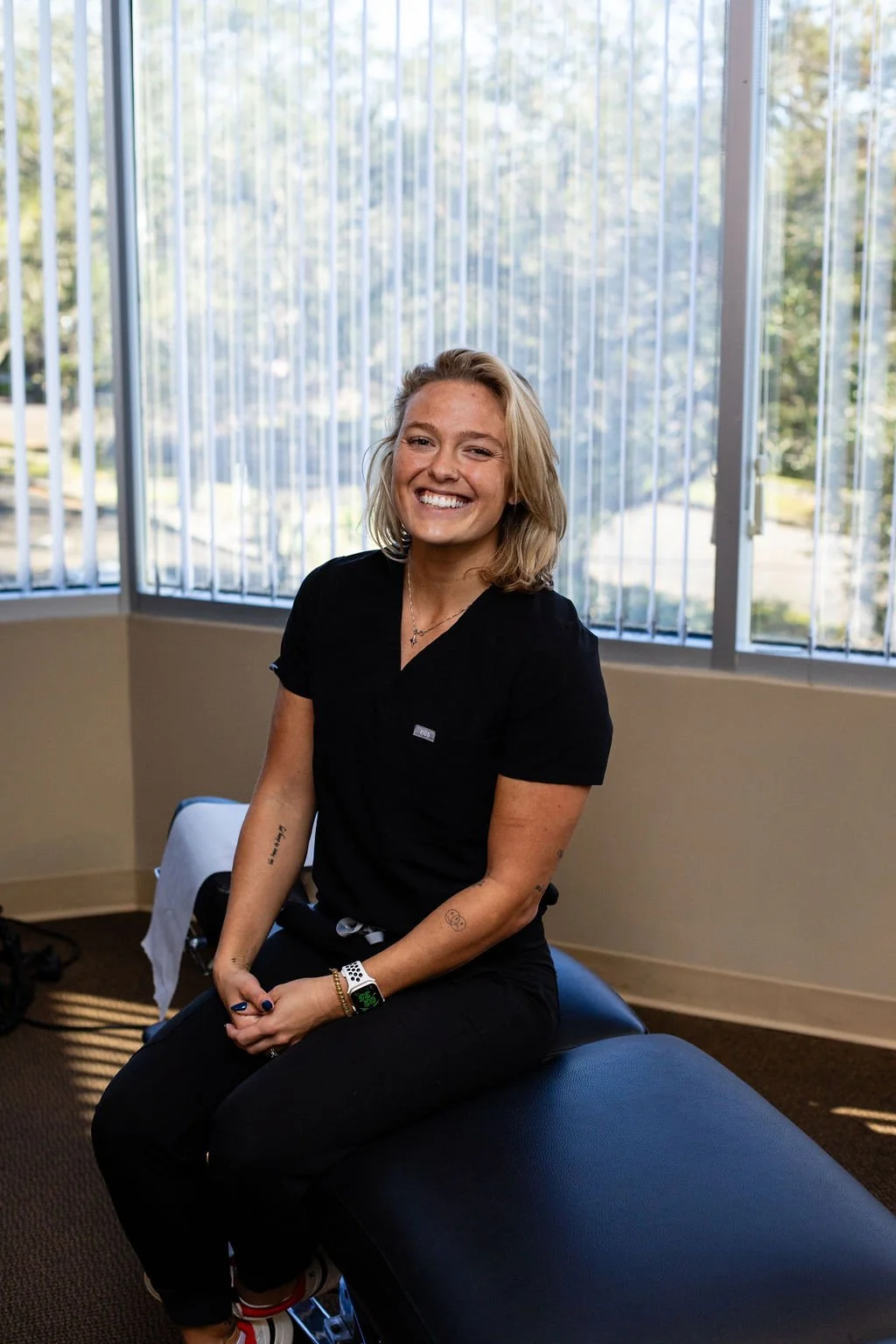 A woman with blonde hair smiling and sitting on a massage table in a room with large windows and vertical blinds.