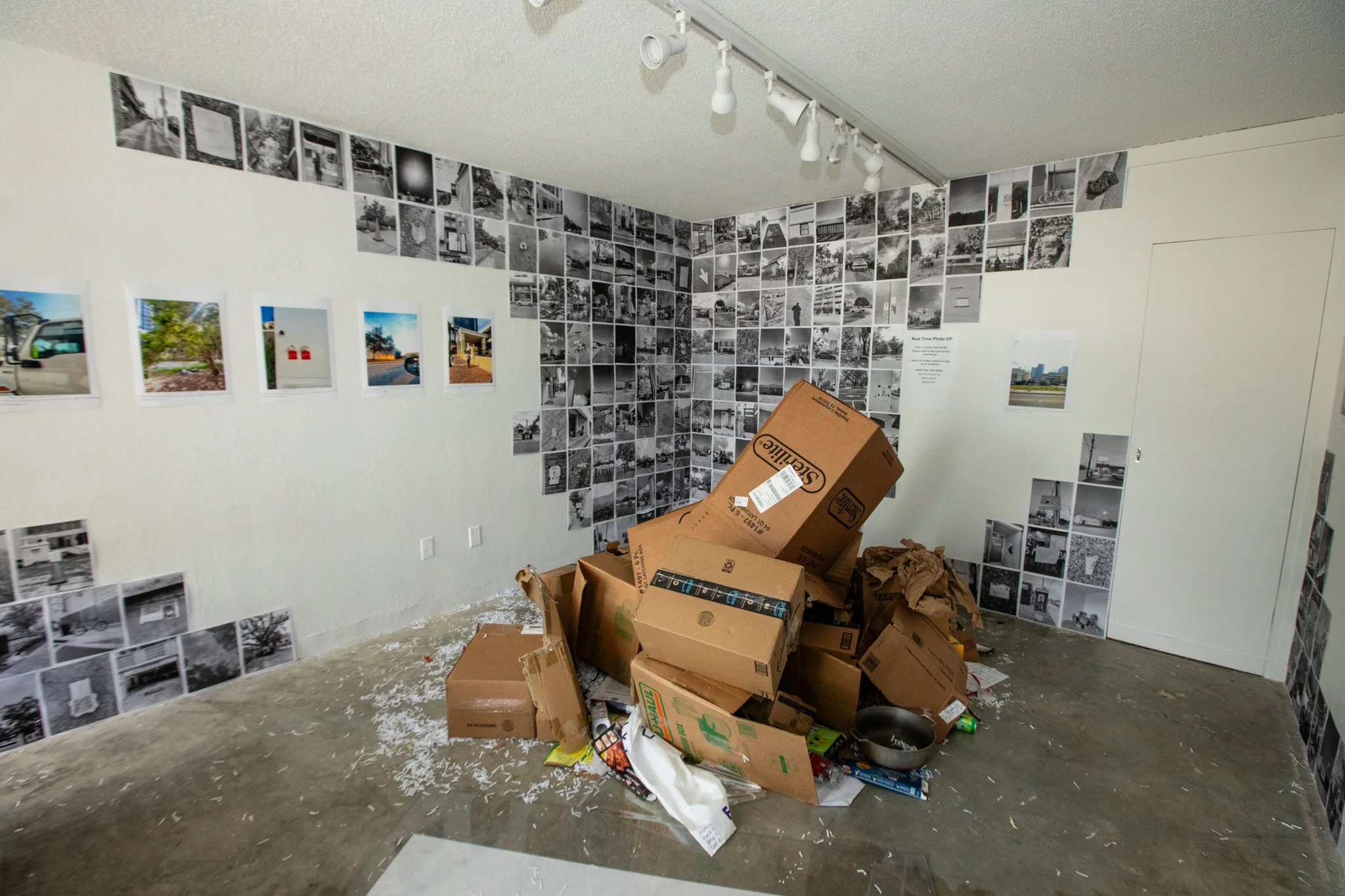 An art gallery with black-and-white photo collages on the walls, cluttered with cardboard boxes, torn packing material, and miscellaneous items on the floor.