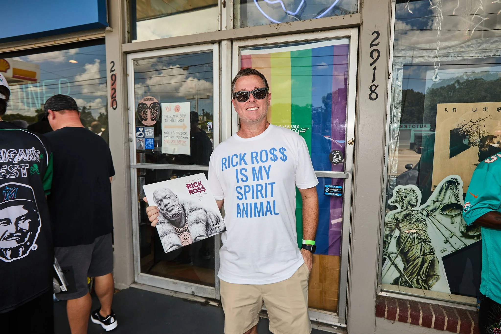 A smiling man wearing sunglasses and a white t-shirt that says 'RICK ROSS IS MY SPIRIT ANIMAL' stands outside a shop holding a magazine featuring Rick Ross. There are people around, and the shop window displays a rainbow flag and various posters.