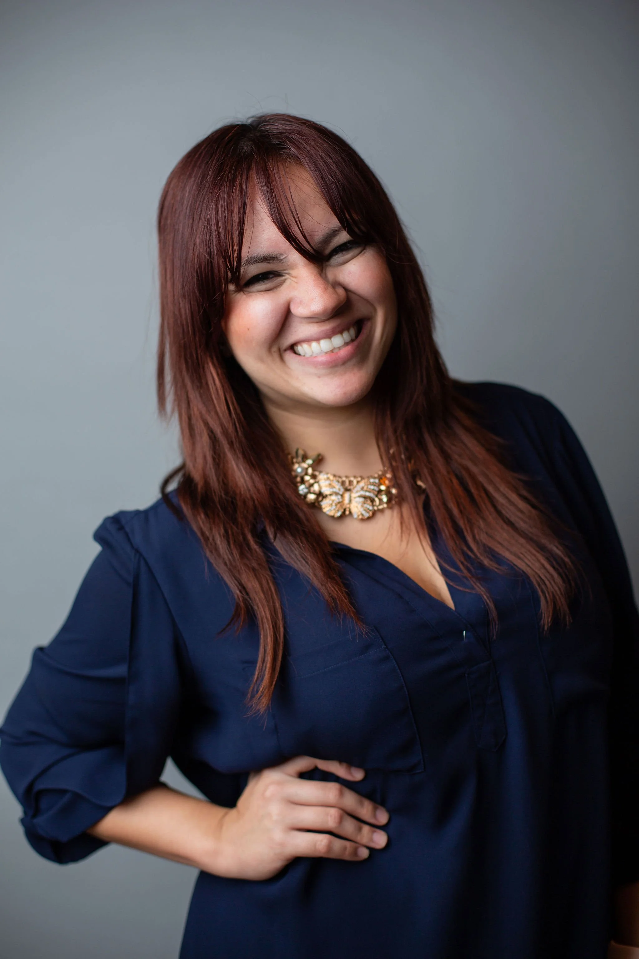 Smiling woman with red hair, wearing a navy blue shirt and a decorative necklace, posing with one hand on her hip against a plain gray background.