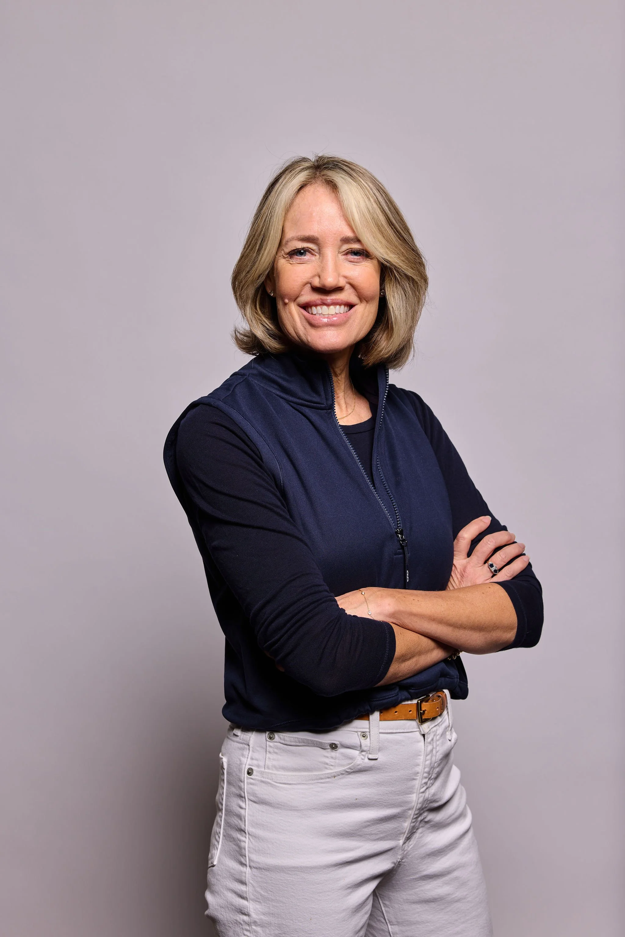A woman with shoulder-length blonde hair smiling, standing with arms crossed against a plain gray background, wearing a navy zip-up jacket and white jeans.