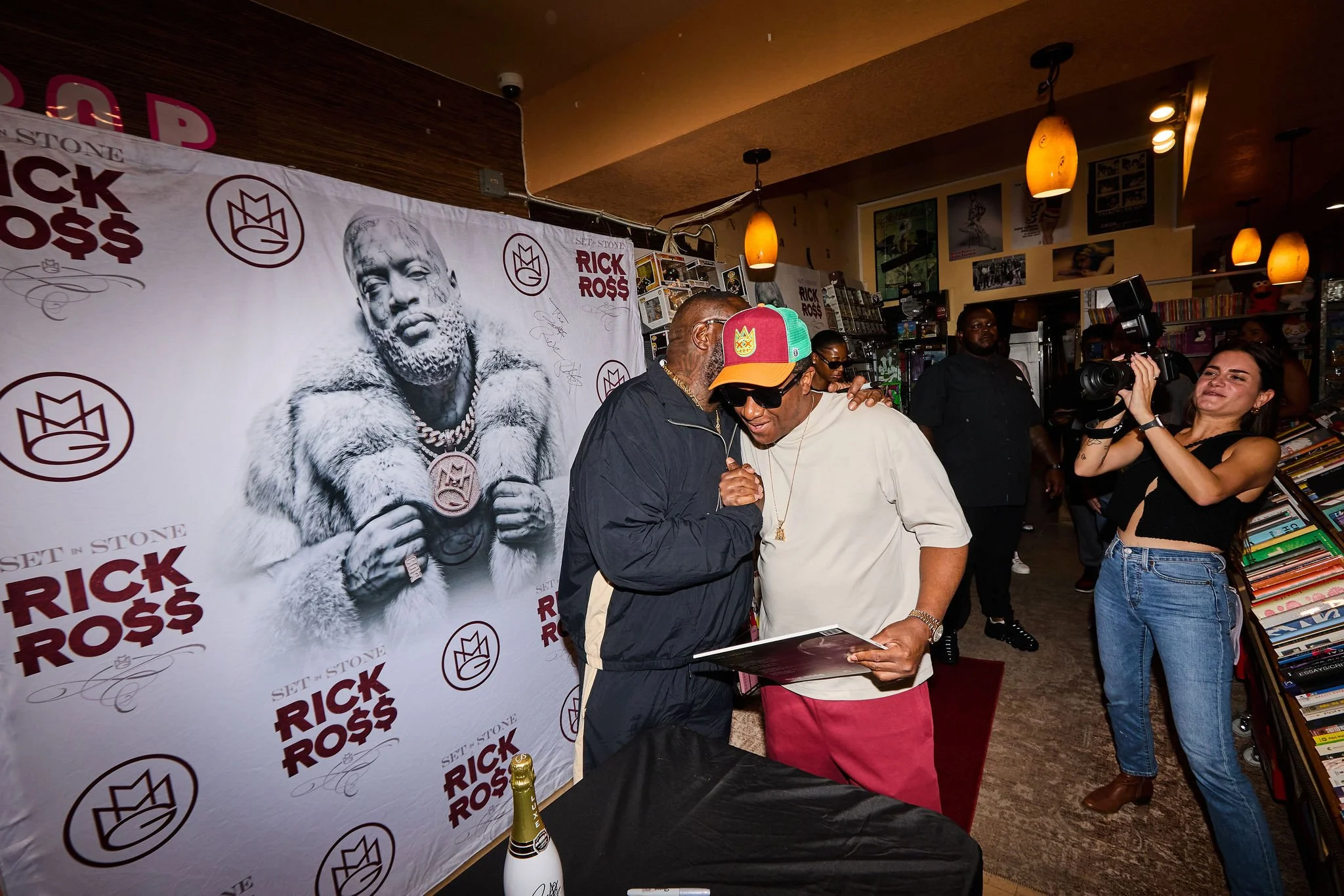 People at a book signing event with a Rick Ross poster in the background, books on display, and a person with a camera recording.