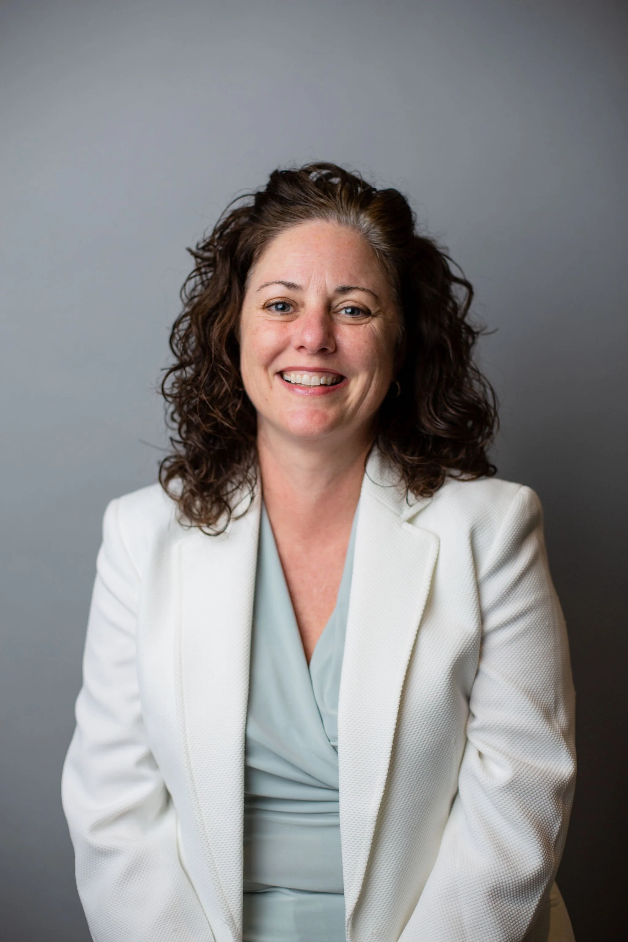 A woman with curly brown hair, blue eyes, and fair skin, wearing a white blazer and light blue blouse, smiling against a gray background.