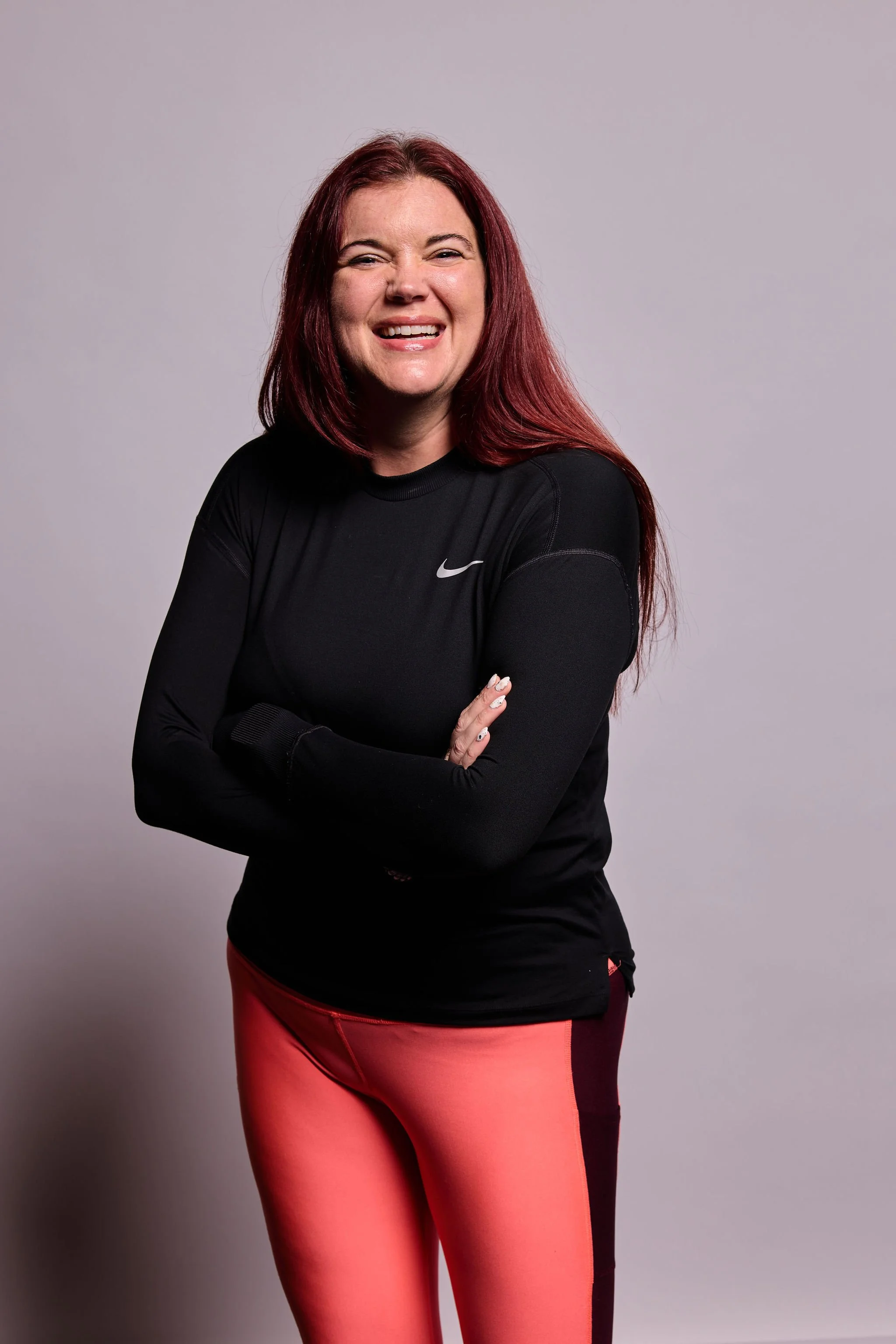 A woman with long red hair, wearing a black Nike long-sleeve shirt and pink leggings, standing against a gray background. She is smiling with crossed arms.