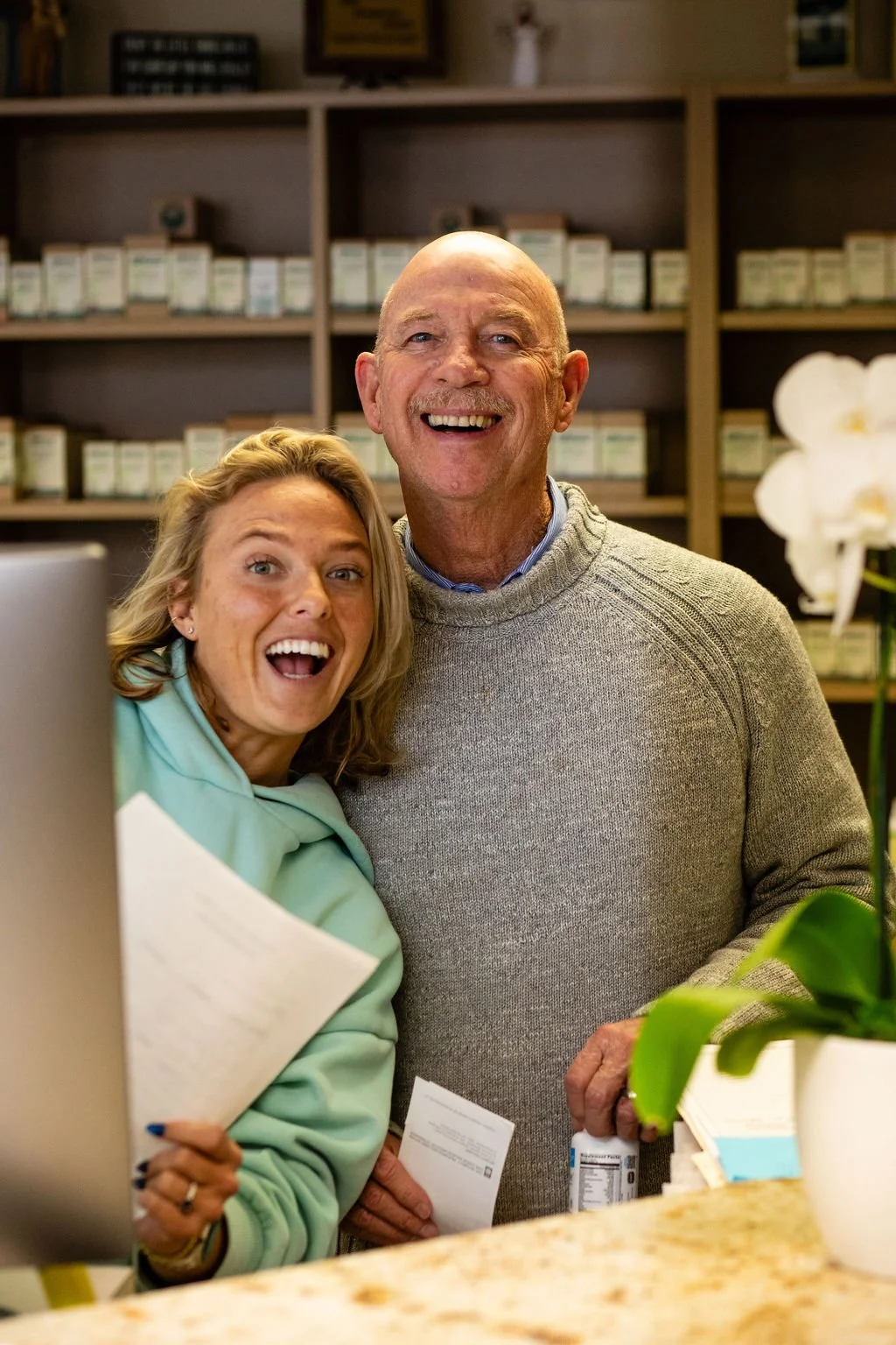A woman and an older man smiling happily at a counter in what appears to be a pharmacy or store, with shelves of medication in the background and a white orchid plant nearby.