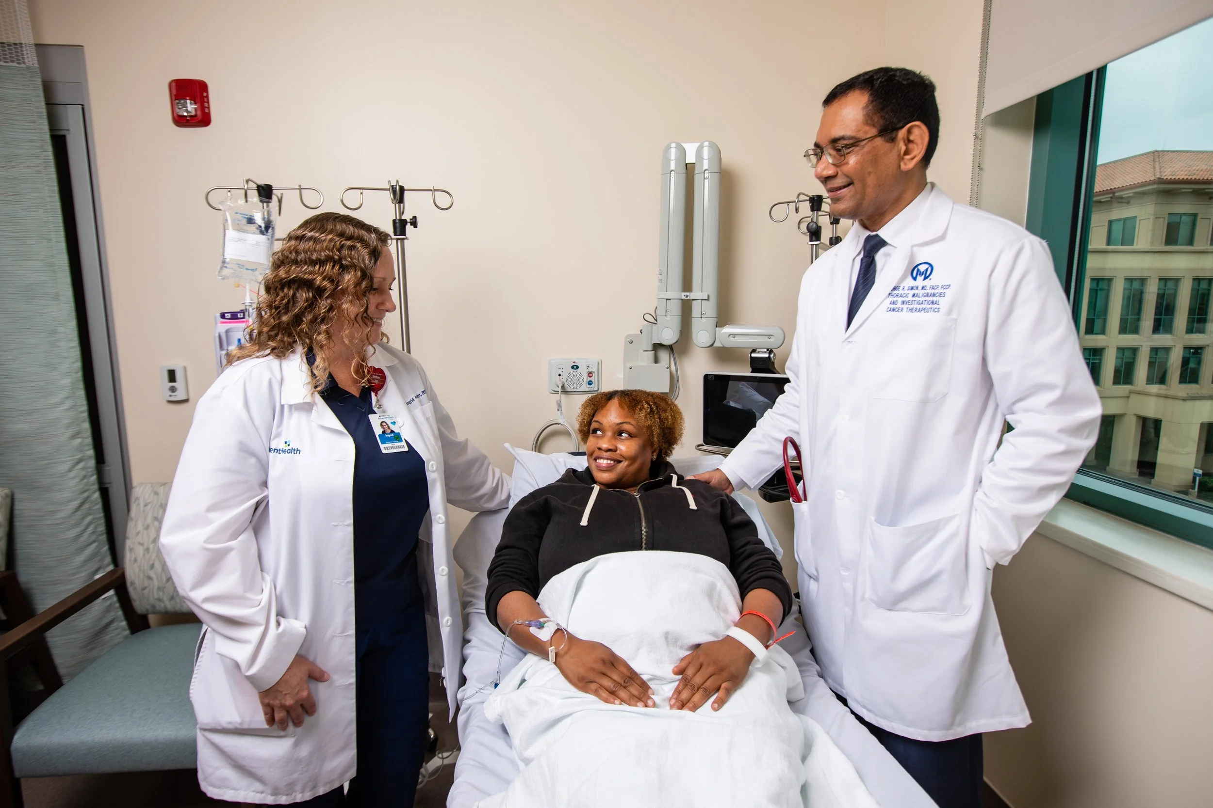 Medical staff, including a nurse and two doctors, visiting an African American woman patient in a hospital room with medical equipment and a large window.