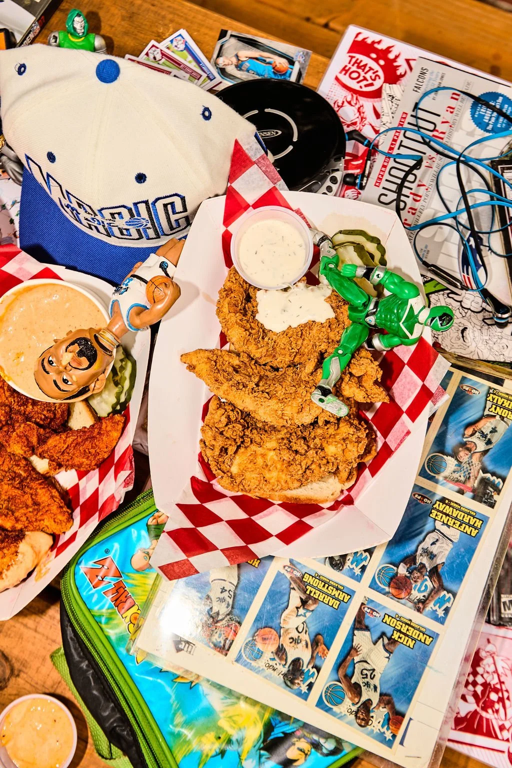 Fried chicken with dipping sauce, a green toy figure, a mug of beer, and various toys and sports memorabilia on a table with a basketball-themed paper.