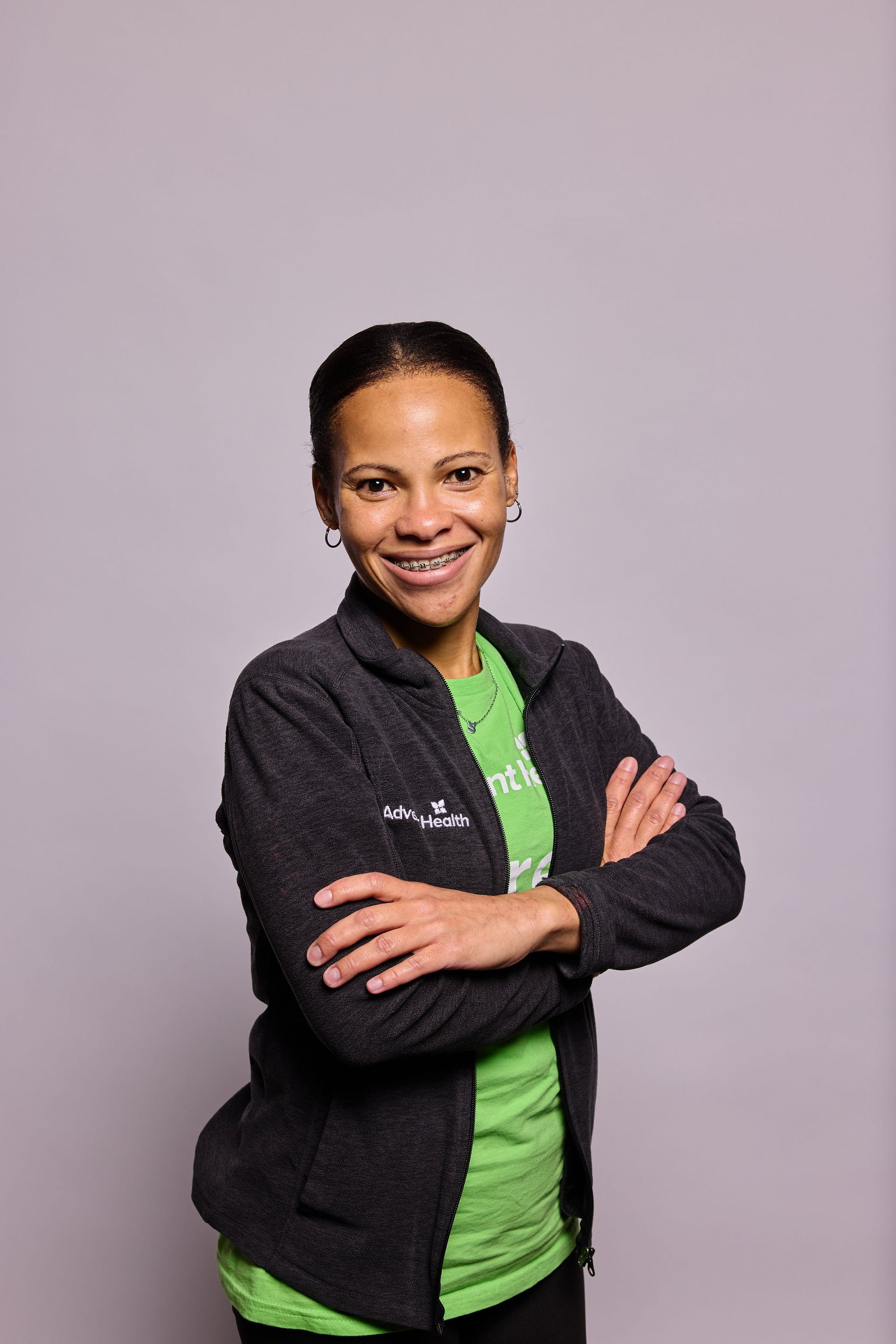 A woman with dark hair in a ponytail, wearing a black jacket with a logo, a green shirt underneath, and sporting braces, stands with arms crossed and smiling in front of a plain gray background.