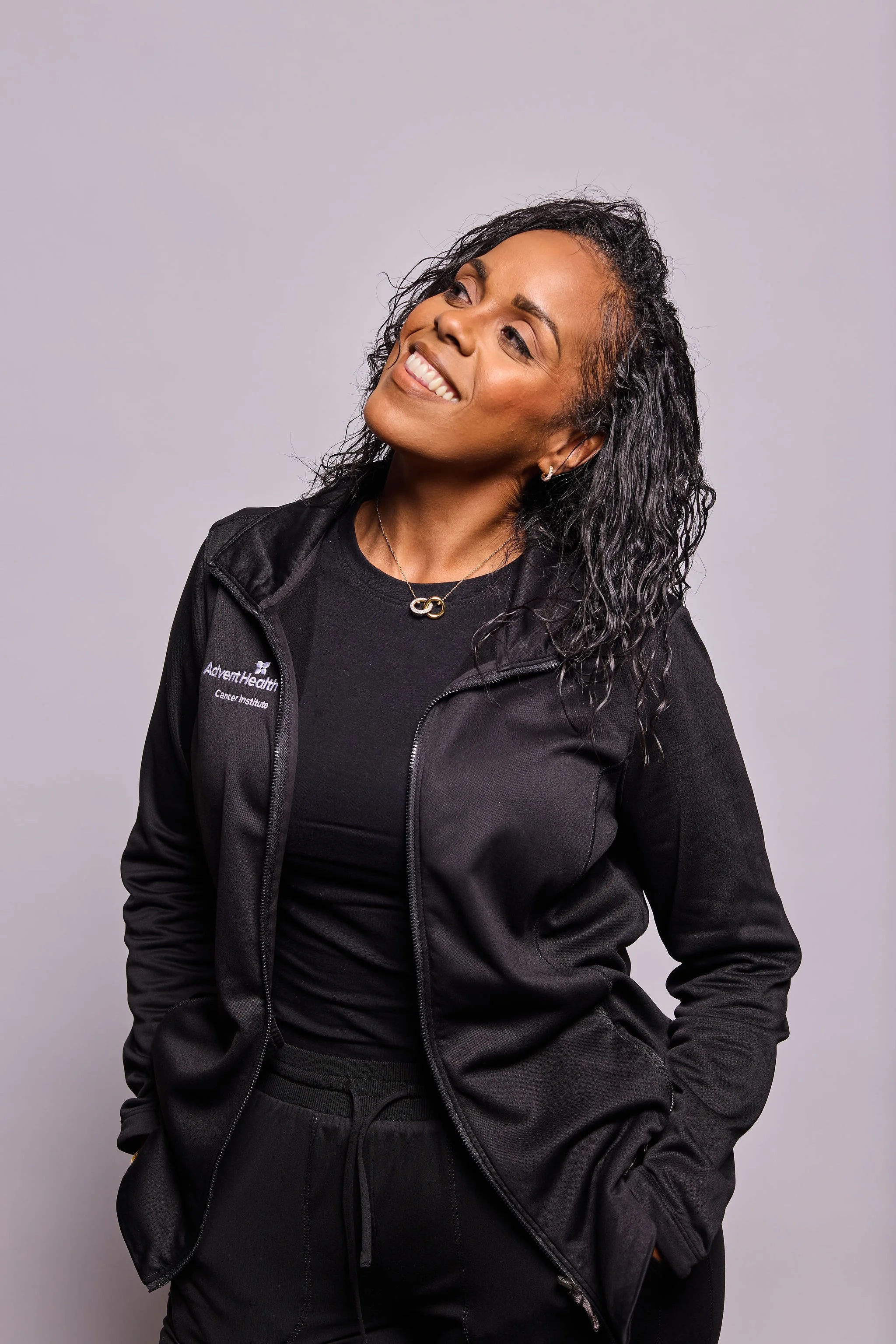 A woman smiling, wearing a black Athletic Health jacket, a black shirt, a necklace with an infinity symbol, and earrings, standing against a light purple background.