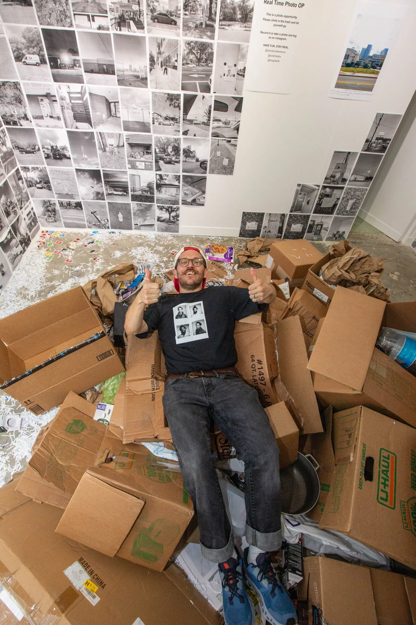 A man lying in a cardboard box surrounded by scattered cardboard boxes and paper debris, giving a thumbs-up and smiling. Behind him are black and white photographs on the wall.