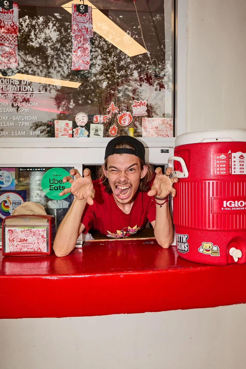 A young man making a playful, fierce face behind a fast food counter with a large red drink cooler on the right and window decals behind him.