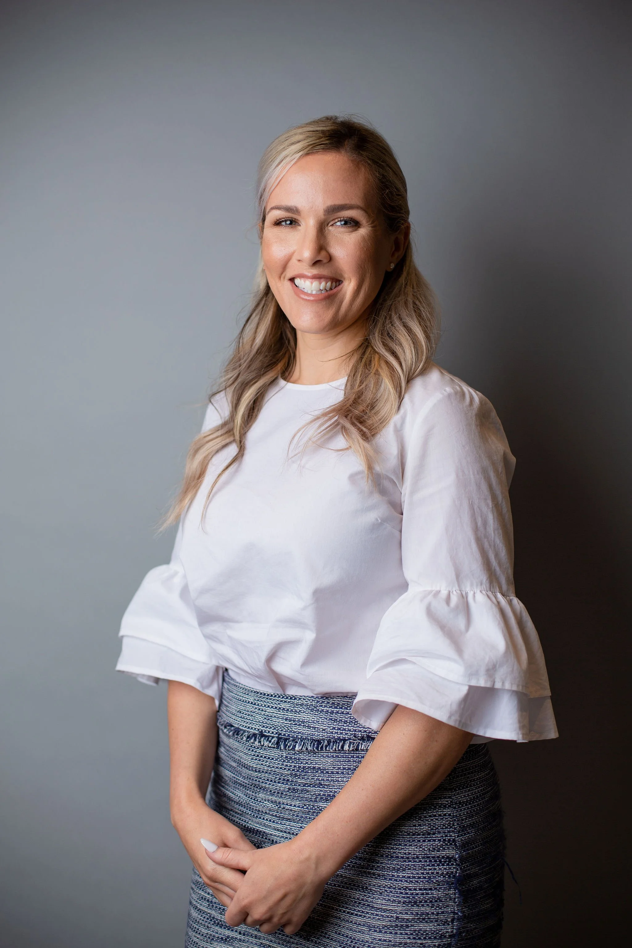 A woman with blonde hair wearing a white ruffled blouse and a patterned skirt, smiling and standing against a grey background.
