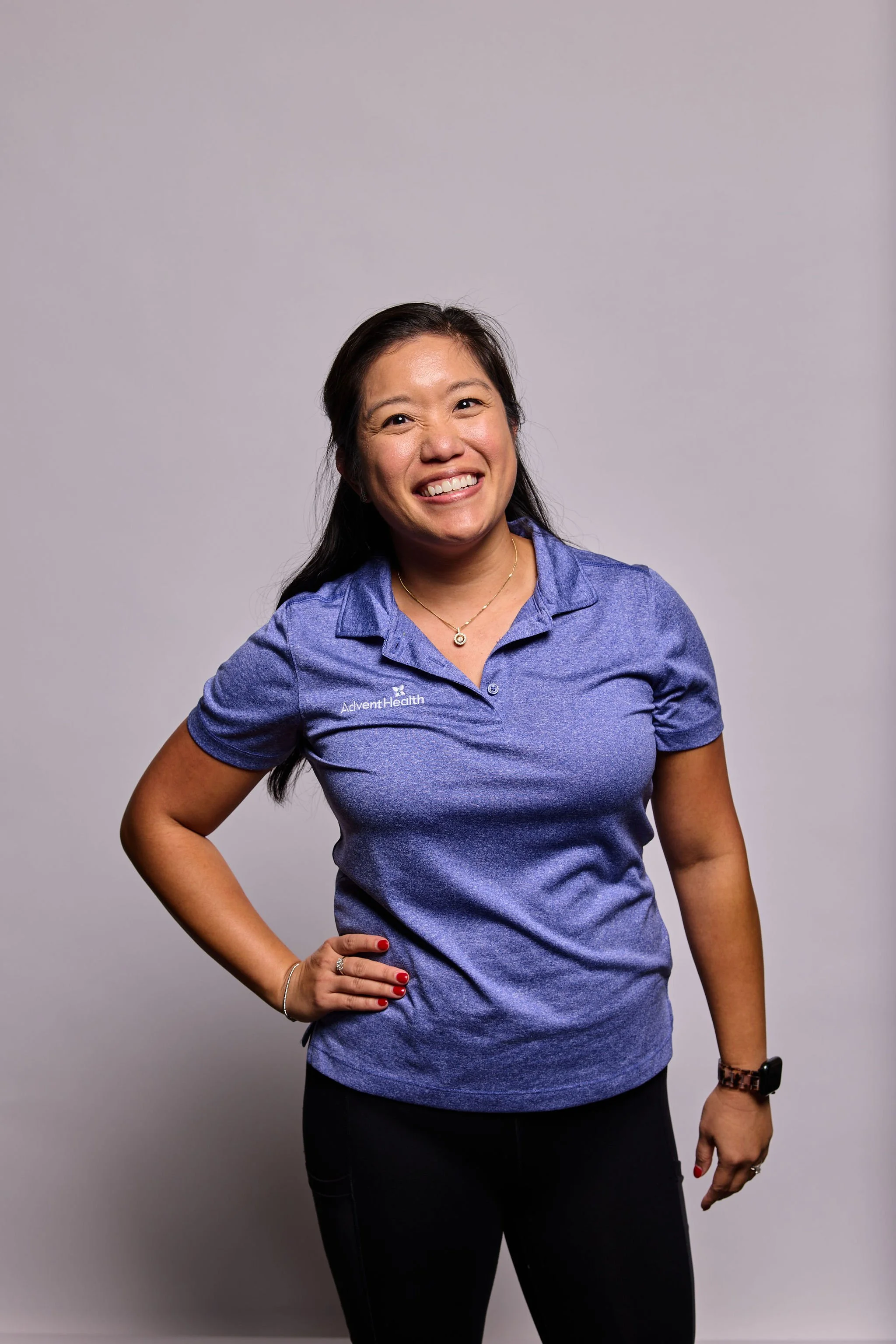 A woman with medium skin tone and long dark hair, smiling and posing with her right hand on her hip, wearing a blue polo shirt with the logo 'AdventHealth' and black pants, standing against a plain gray background.