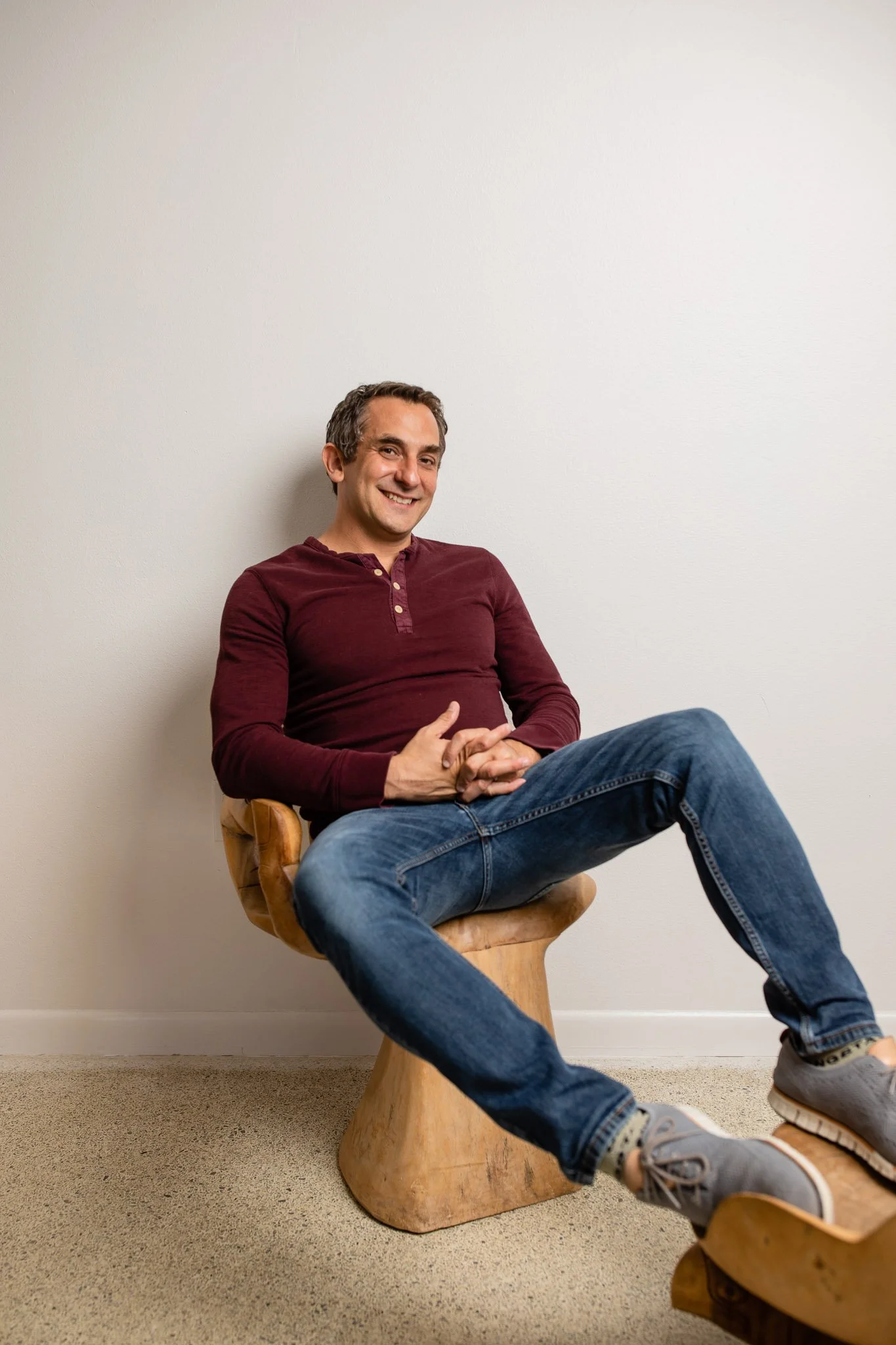 A man sitting comfortably on a wooden chair with his legs crossed and smiling at the camera, against a plain white wall.