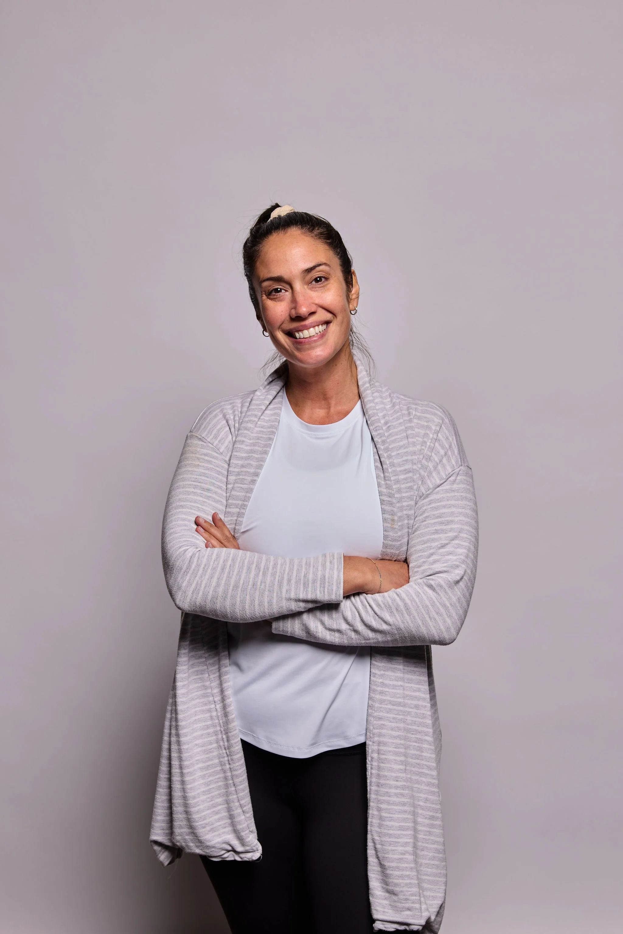 A woman with dark hair tied in a high ponytail, wearing a white shirt, gray cardigan, and black pants, smiling with her arms crossed, standing in front of a plain gray background.