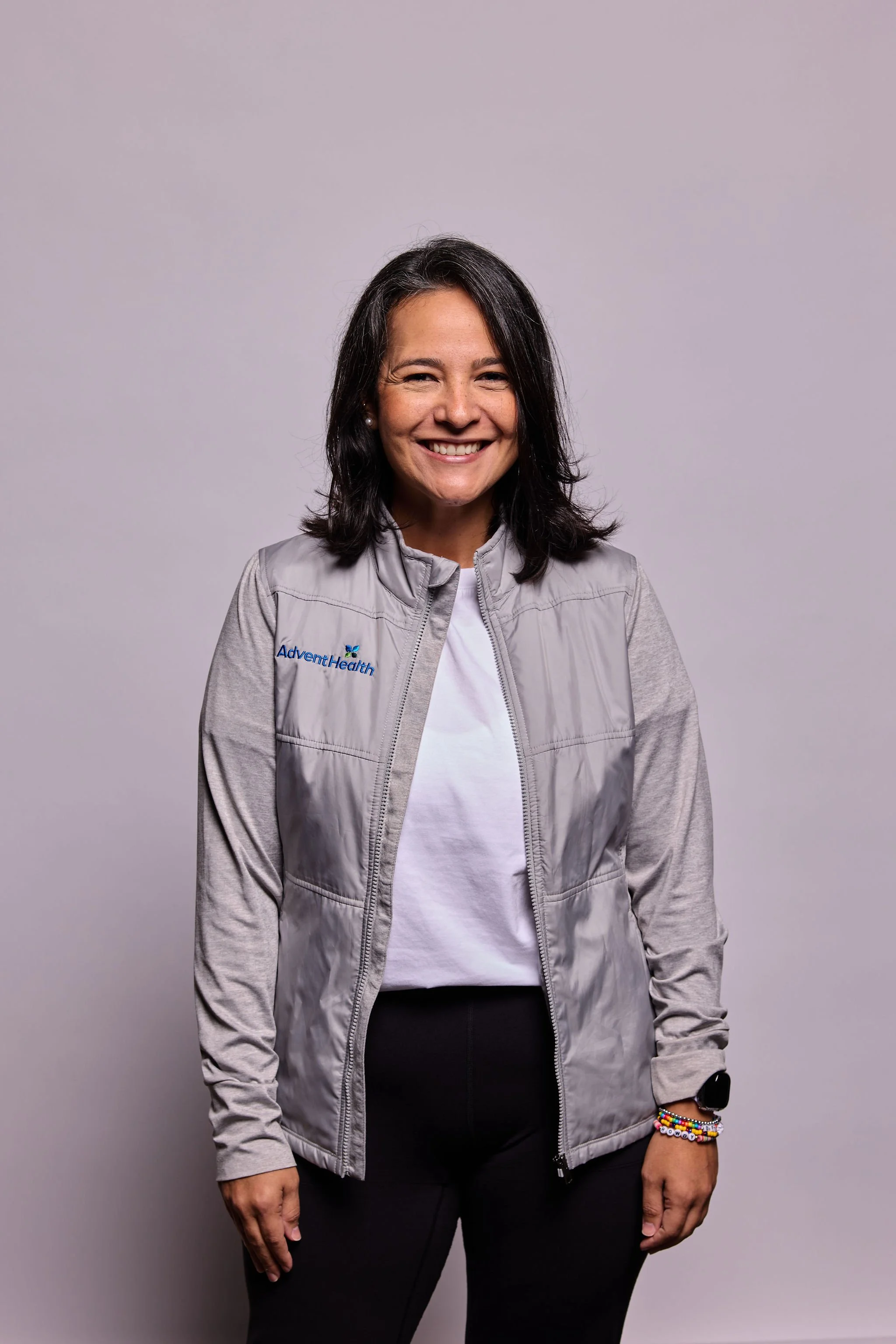 A smiling woman with dark hair wearing a light gray jacket with 'AdventHealth' logo, a white shirt, black pants, and colorful beaded bracelets on her left wrist, standing against a plain gray background.