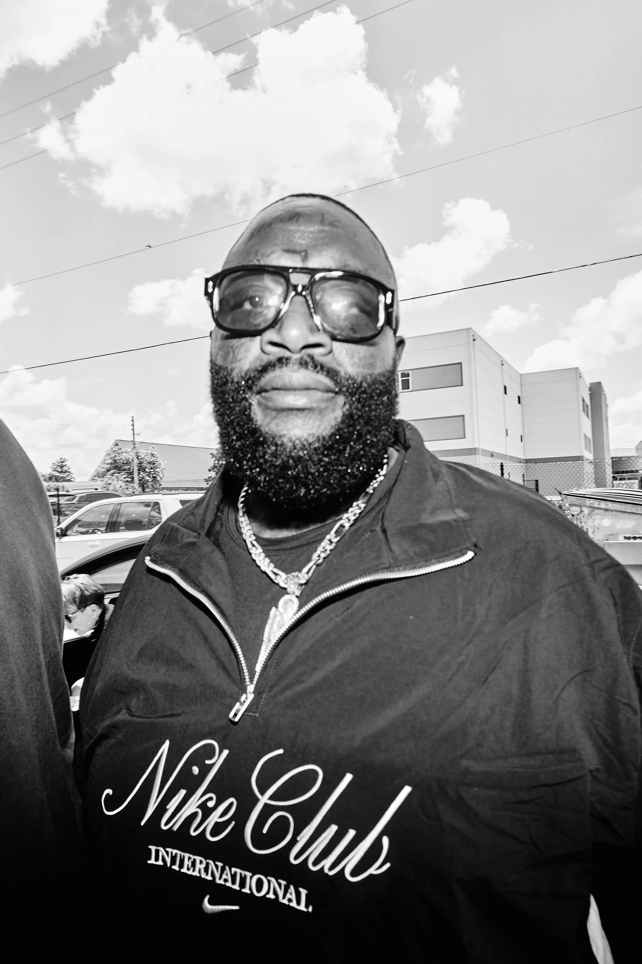 Close-up black-and-white photo of a man wearing large glasses, a chain necklace, and a Nike Club shirt, standing outdoors in a parking lot with modern buildings, trees, and a partly cloudy sky in the background.