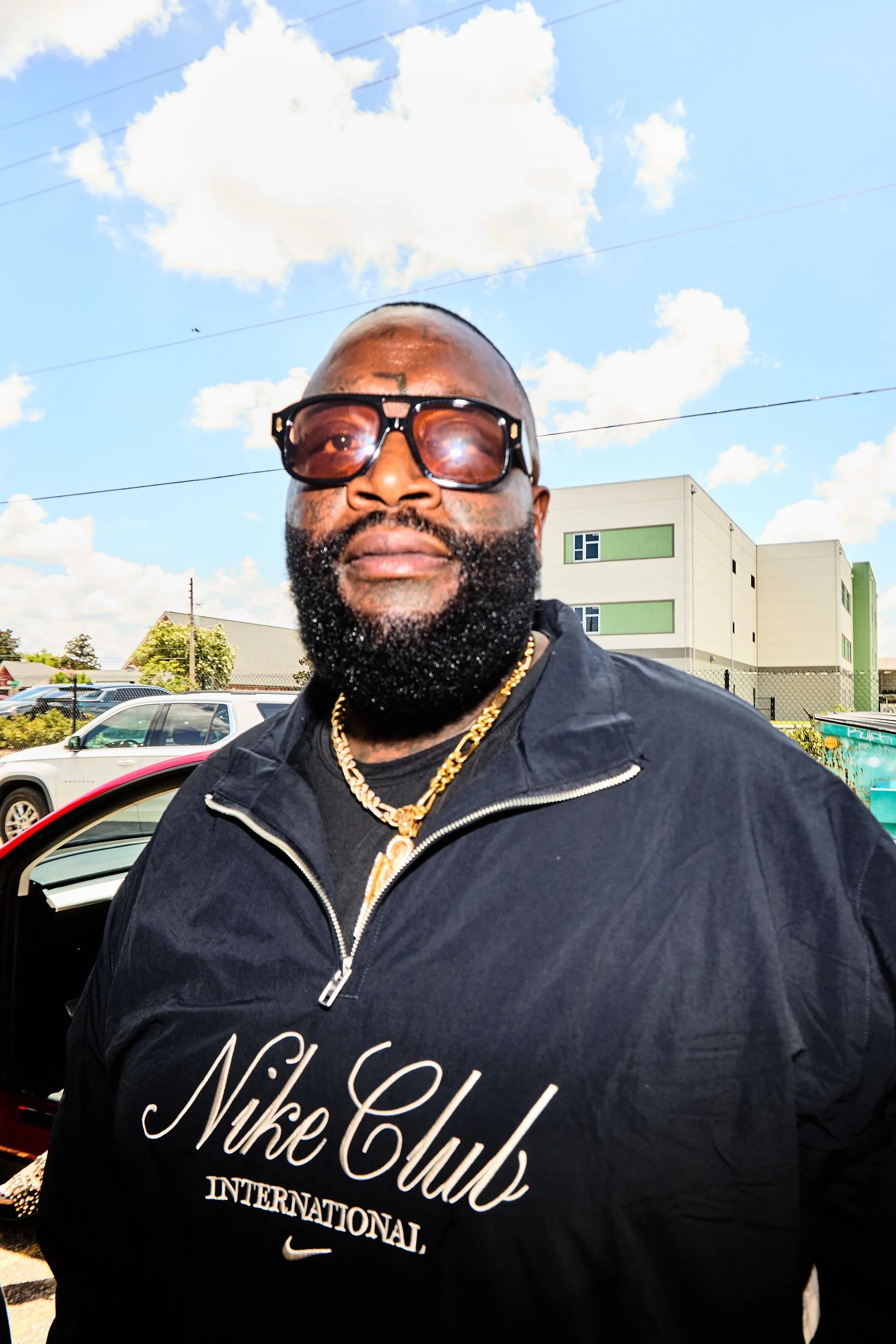 A man with a beard, wearing sunglasses, gold chains, and a black Nike club jacket, standing outdoors in a parking lot with cars and a multi-story building in the background, under a partly cloudy sky.