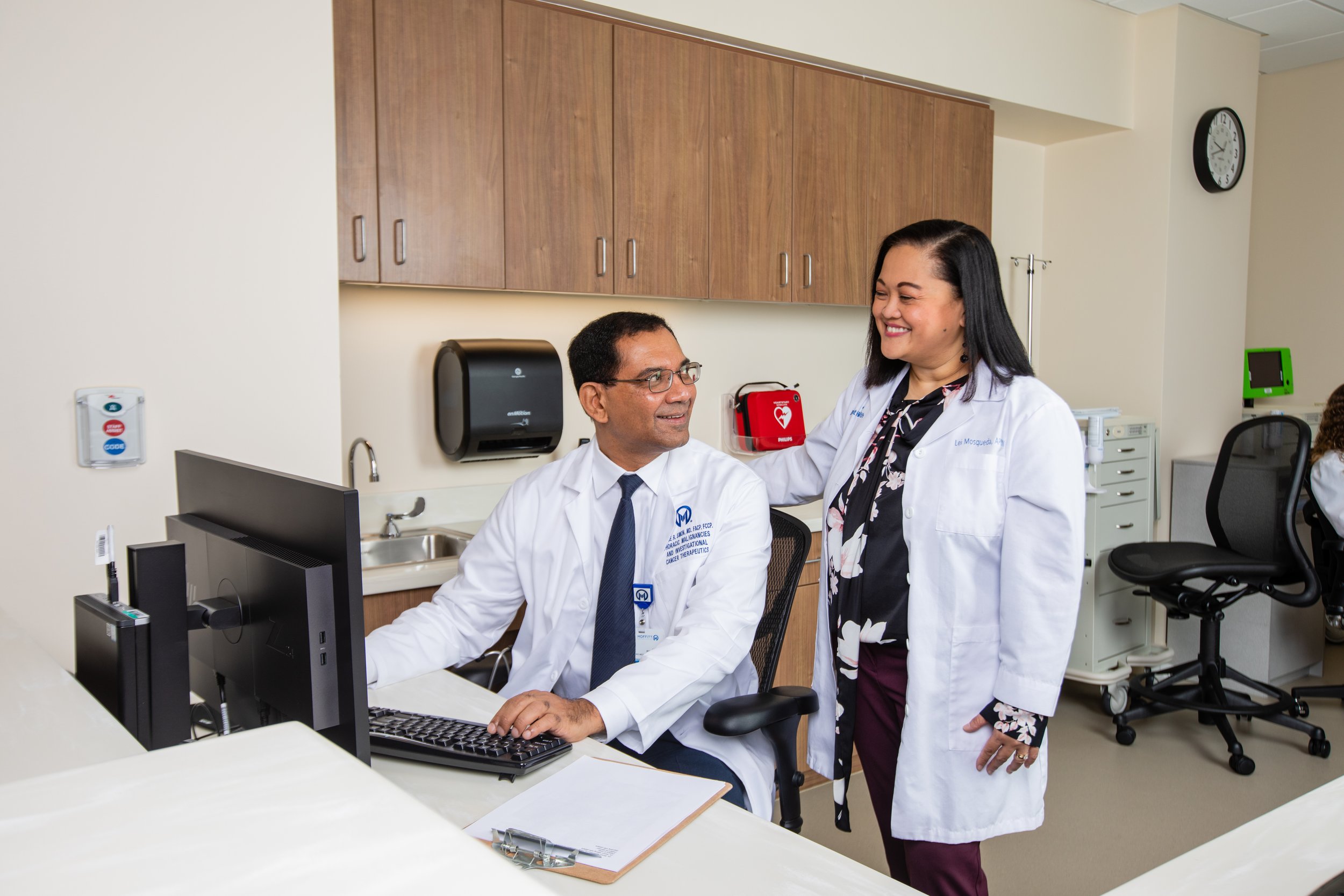 A male doctor sitting at a desk in a hospital room, smiling at a female nurse standing beside him with her hand on his shoulder, both wearing white lab coats.