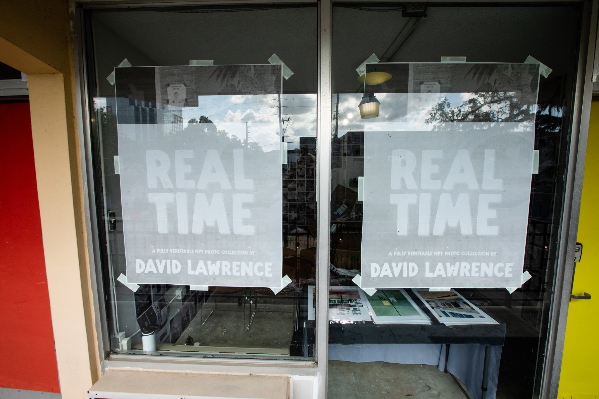 Storefront with two large white posters on the glass door reading 'REAL TIME' and 'A FULLY VERIFIABLE NFT PHOTO COLLECTION BY DAVID LAWRENCE' taped with white tape.