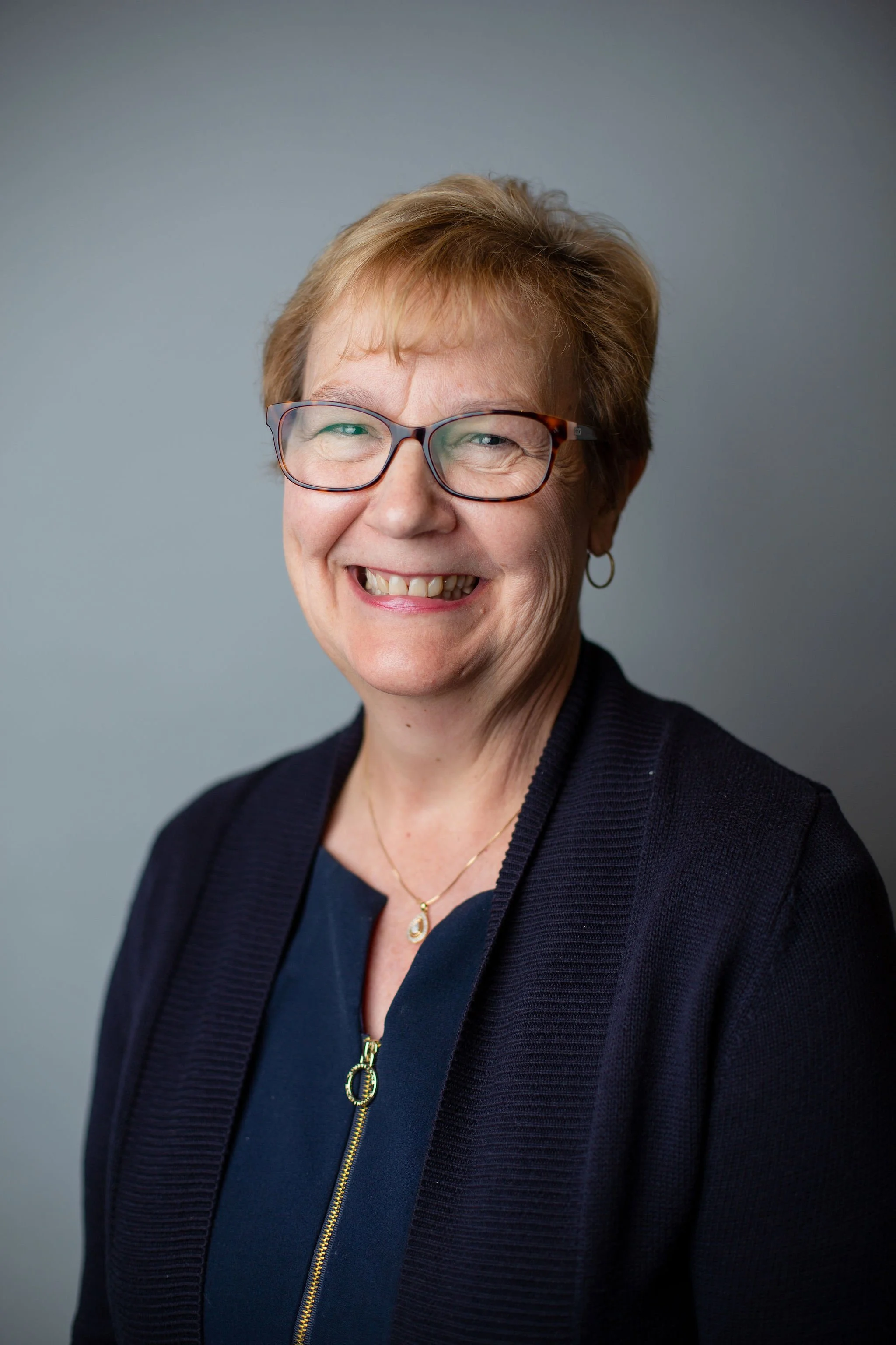 A middle-aged woman with short, light brown hair and glasses smiling at camera, wearing a dark blue top, a navy sweater with a zipper, a gold necklace, and a earring, against a plain gray background.