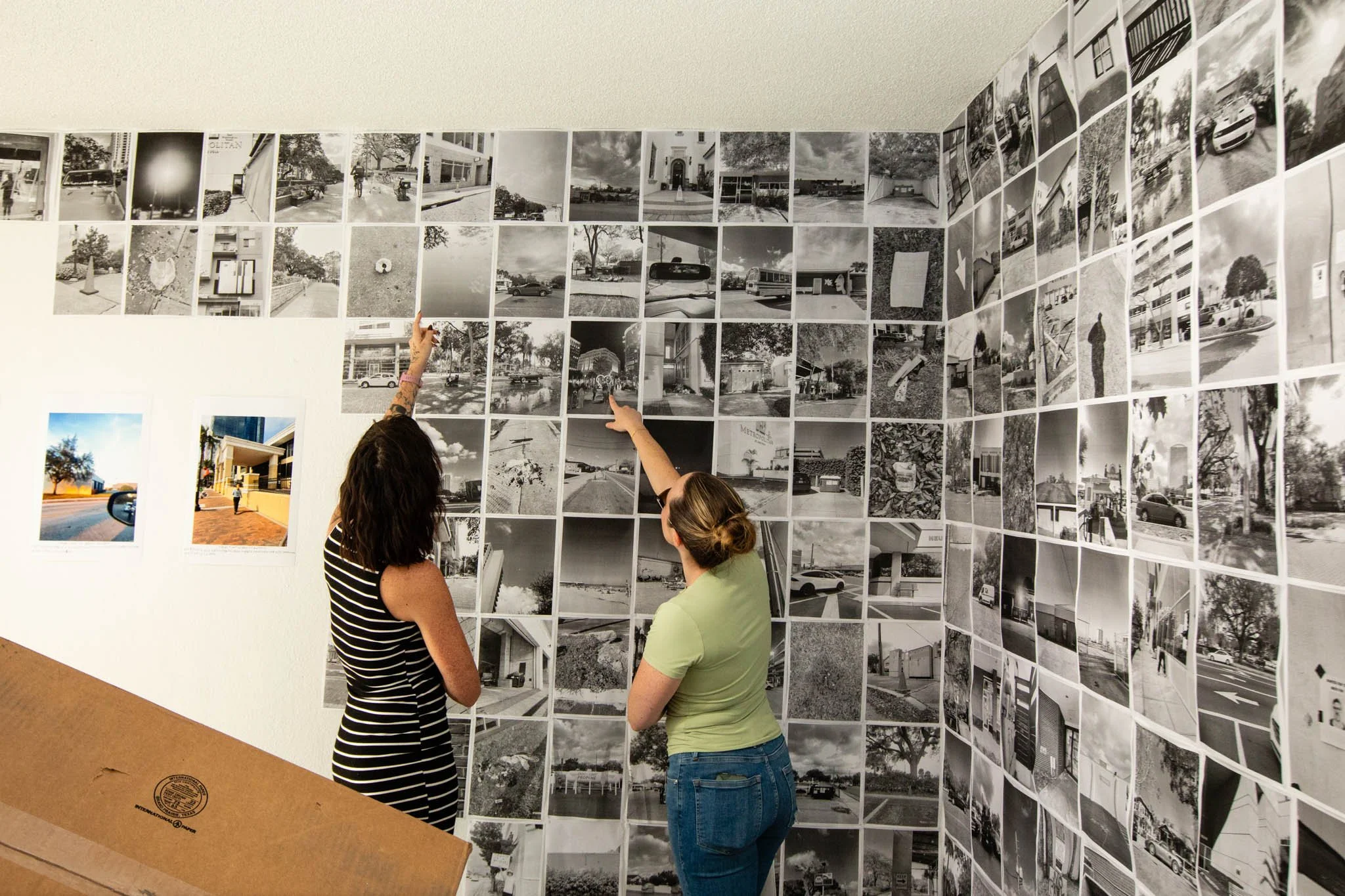 Two women look at a wall covered with numerous black and white photographs of various outdoor scenes in an art exhibit.