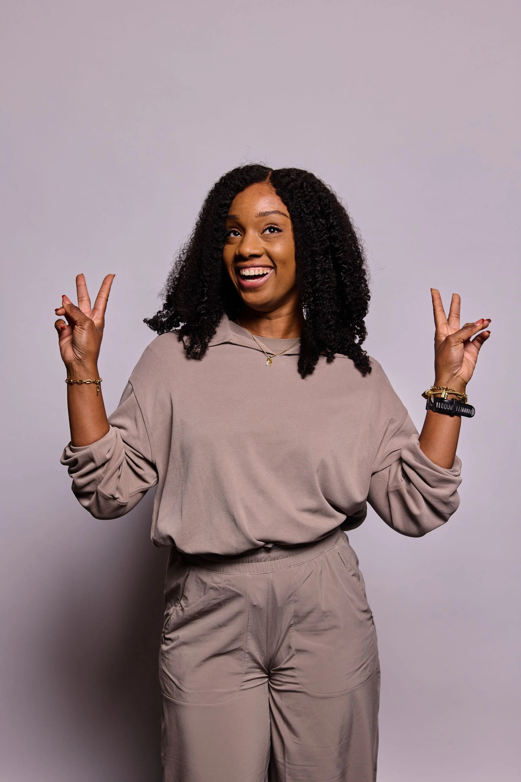 A woman with curly black hair, smiling, making peace signs with both hands, wearing a beige sweatshirt and pants, standing against a plain light gray background.