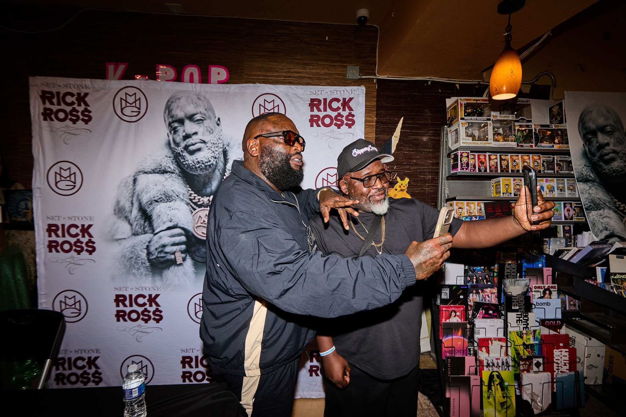 Two men posing for a photo at a Rick Ross promotional event. One man is wearing sunglasses and a black jacket, the other is wearing a black cap and glasses. They are in front of a Rick Ross branded backdrop and a display of Funko Pop figures.