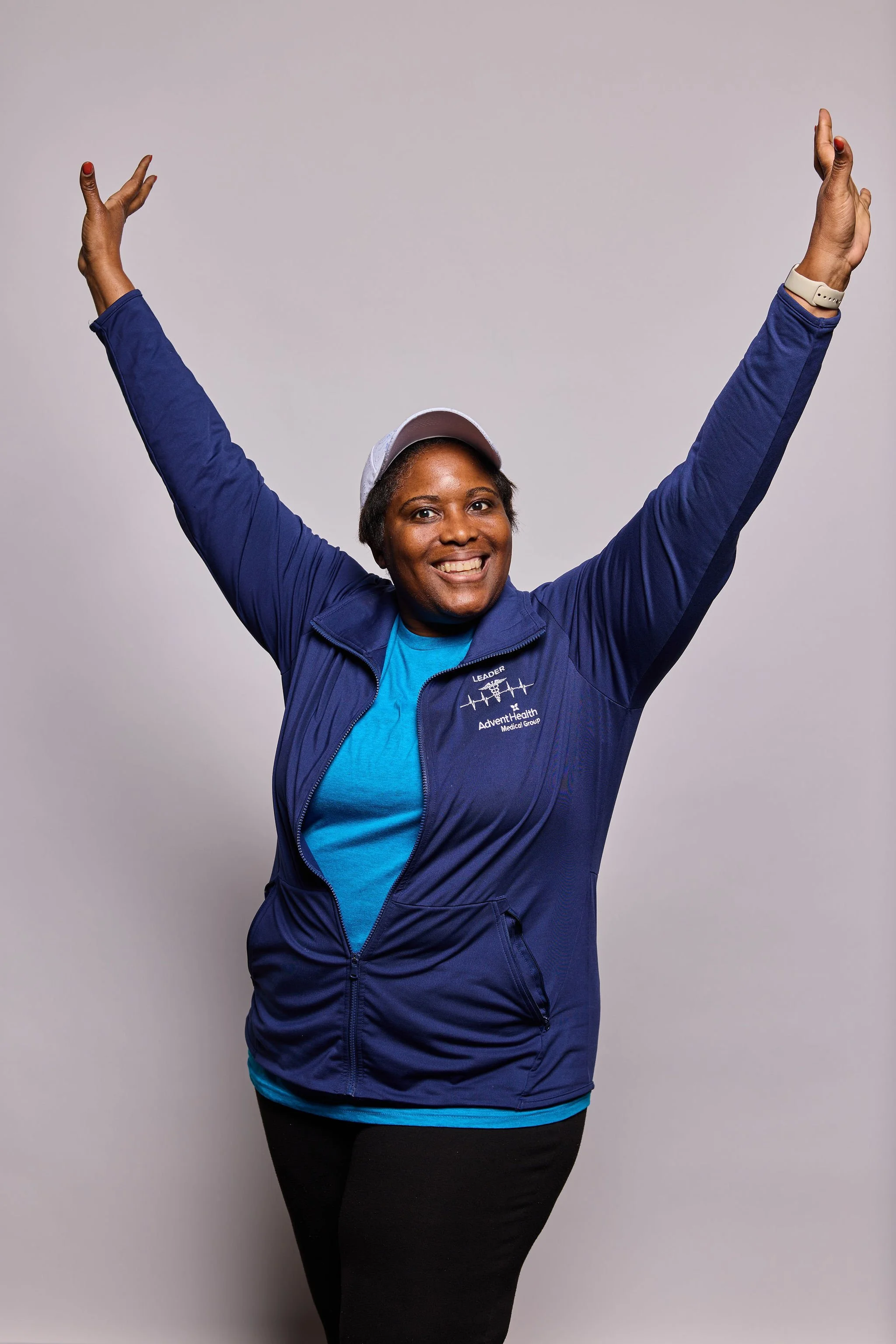 A smiling woman in a blue jacket, black pants, wearing a white cap, with her arms raised in celebration against a plain gray background.