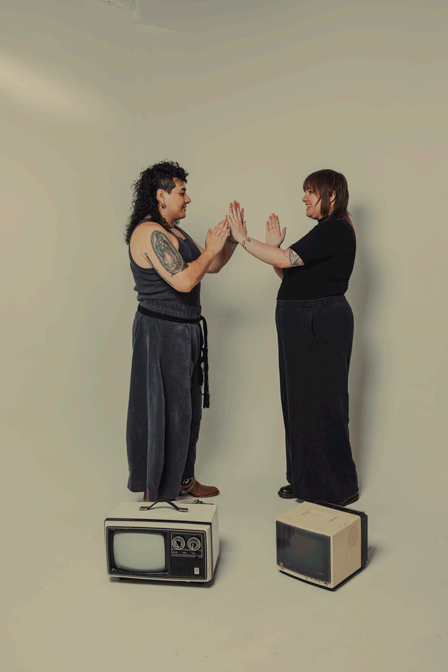 Two women standing on vintage televisions, touching palms and smiling at each other in a neutral setting.