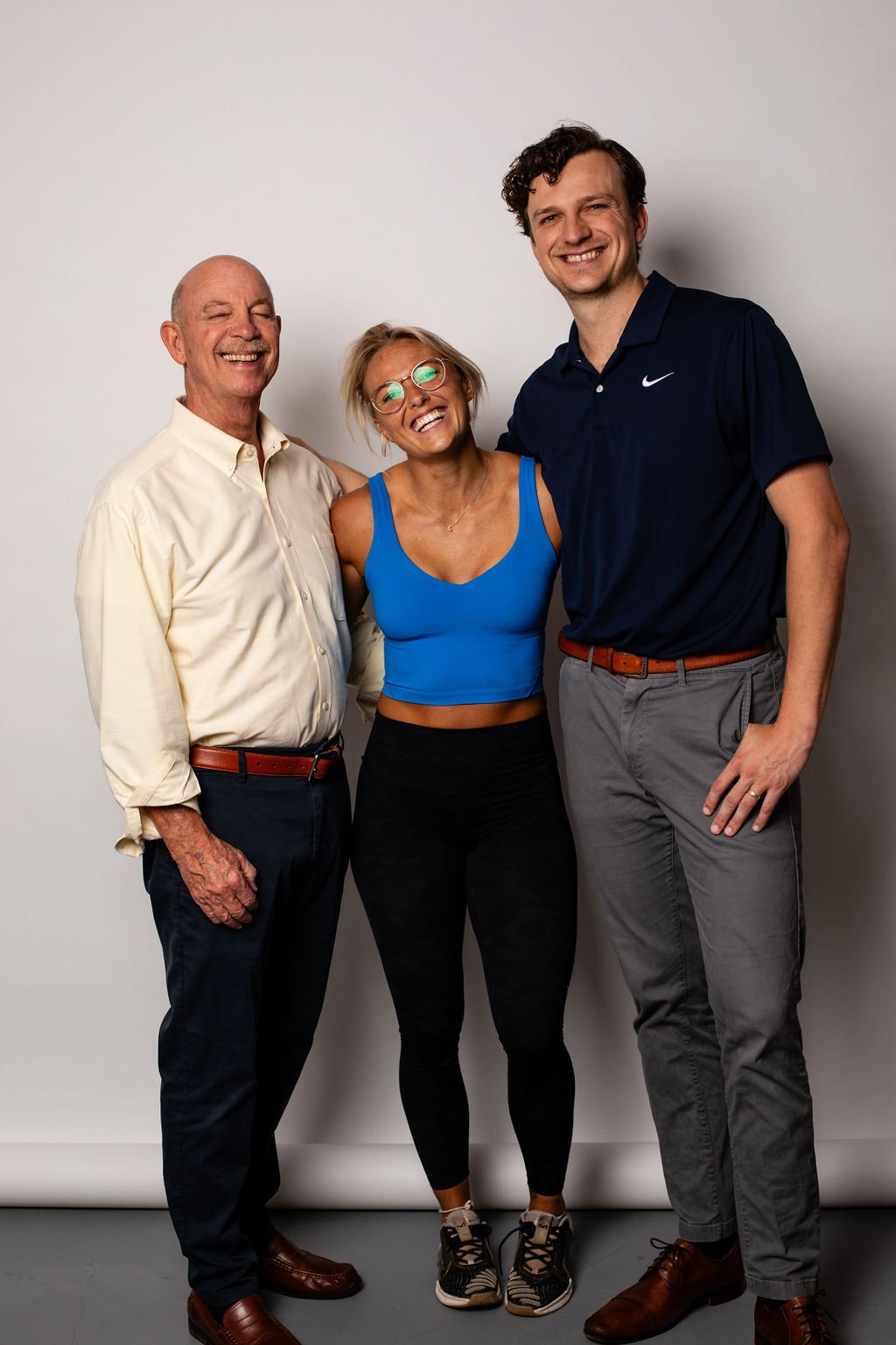 Three smiling people standing together against a plain light-colored background. An older man in a light yellow shirt on the left, a young woman with glasses and a blue sports bra in the middle, and a tall young man in a navy blue polo and gray pants on the right.
