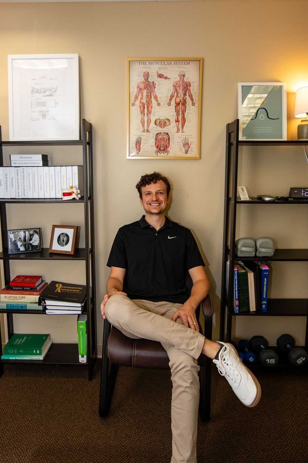 A man sitting on a chair in an office, smiling at the camera. Behind him are two shelves filled with books, framed photos, and medical models. The wall has anatomical posters of the muscular system and a graph.