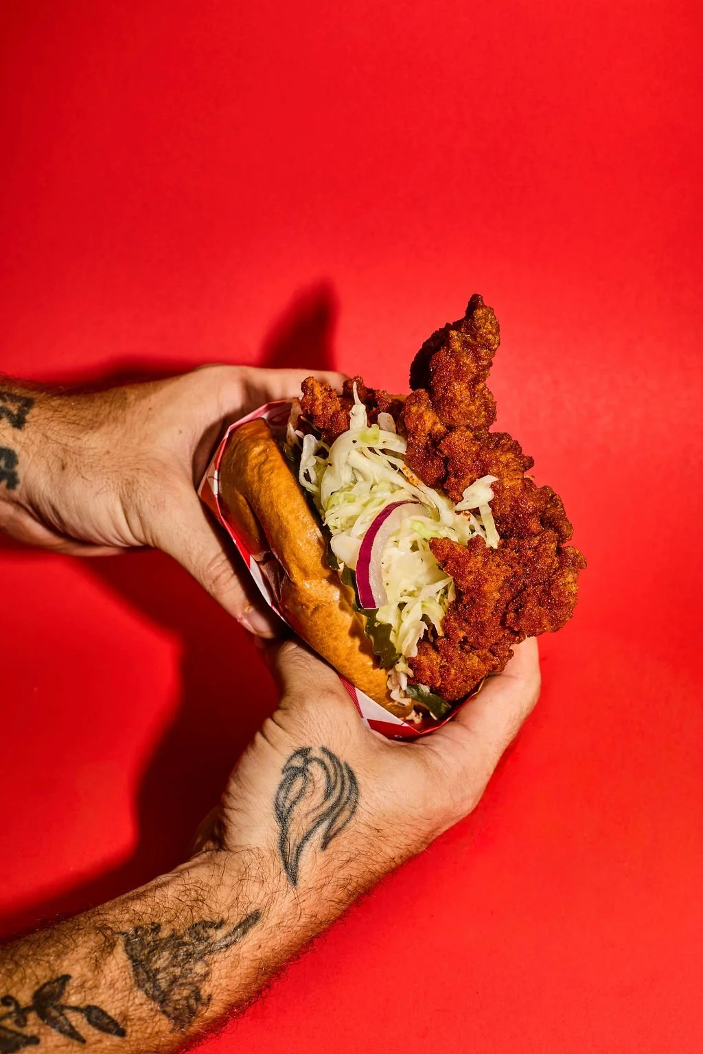 A person with tattoos on both arms holding a fried chicken sandwich with coleslaw, pickles, and onions on a red background.