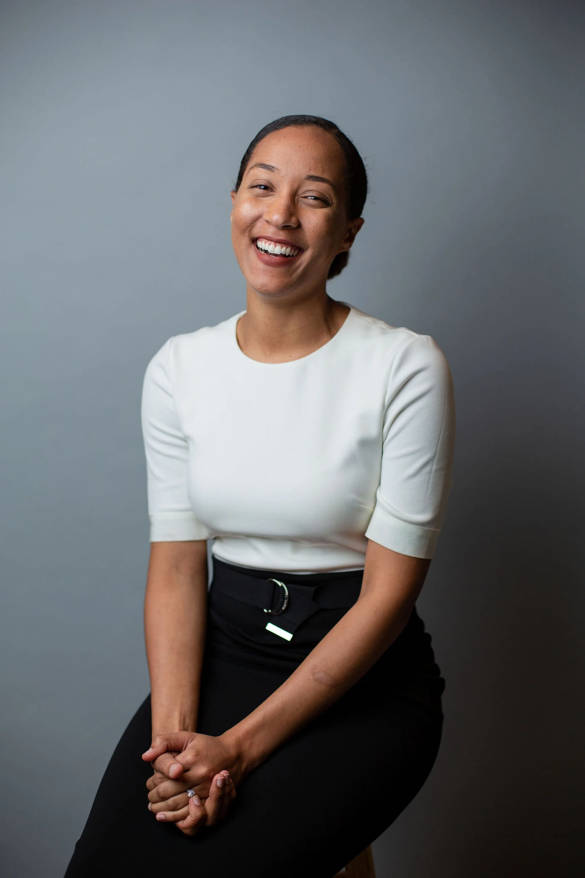 A woman smiling, sitting against a gray background, wearing a white top and black skirt.