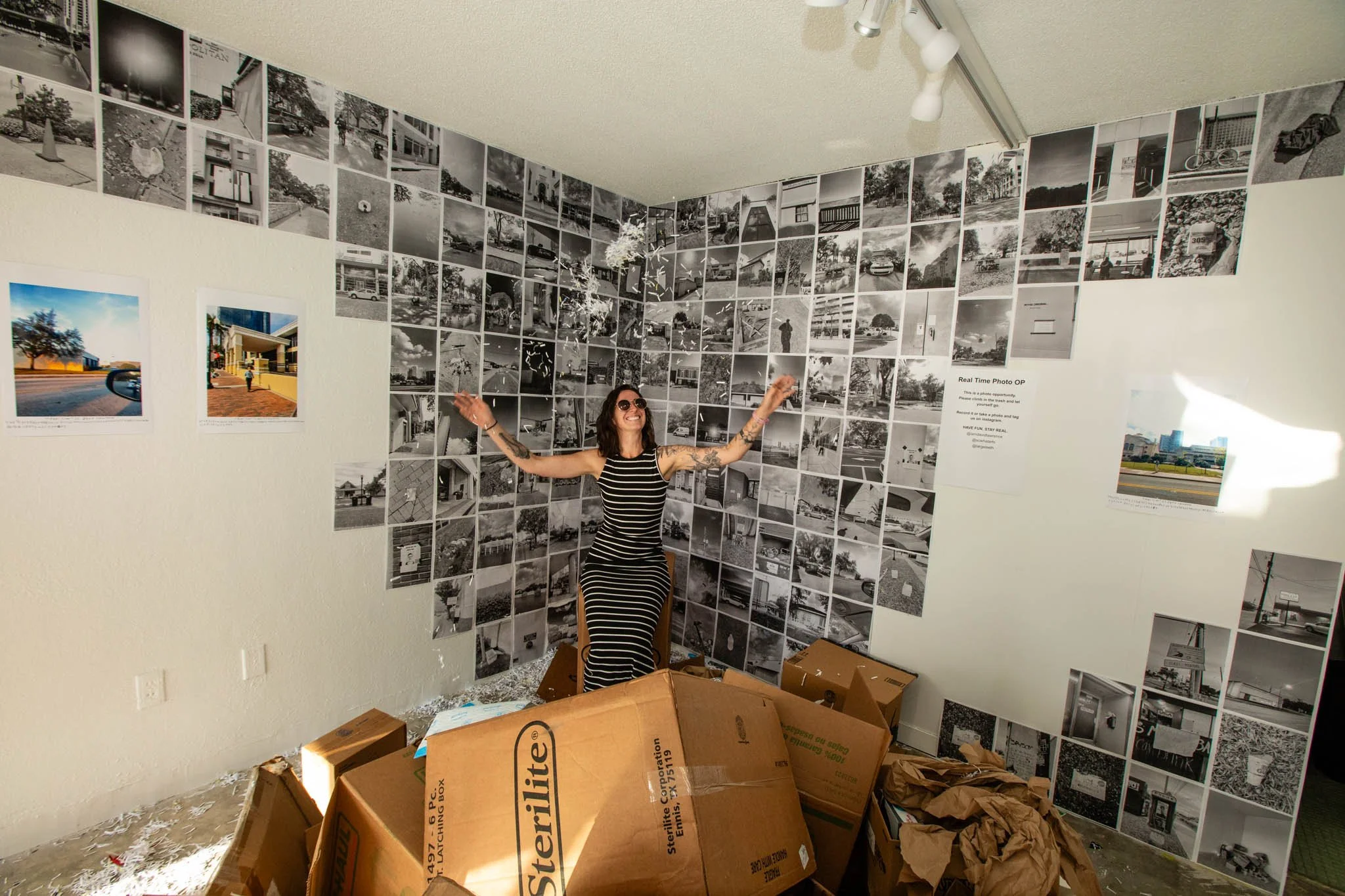 A woman with dark hair, sunglasses, and tattoos on her arms standing with arms outstretched in an art installation featuring a wall covered in black and white small photographs. There are cardboard boxes and crumpled paper on the floor.