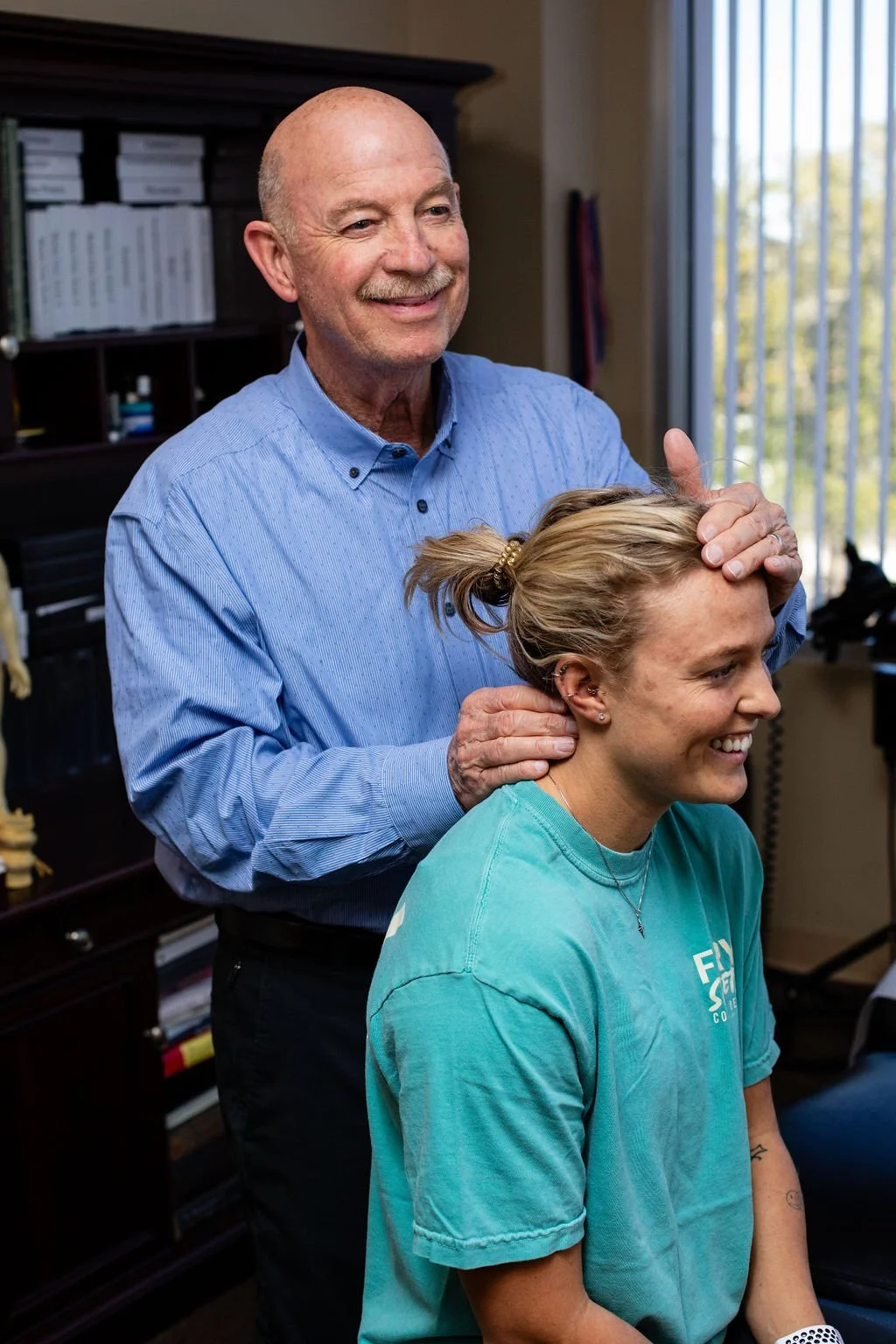 An older man giving a neck massage to a young woman in a teal t-shirt, inside a room with a window and bookshelves in the background.