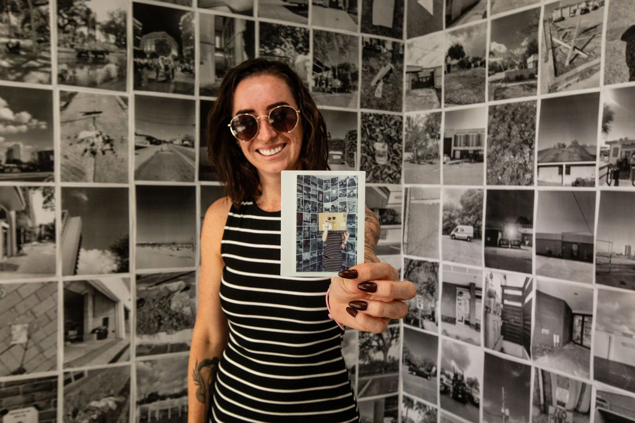 A woman with dark hair, sunglasses, and a striped dress holding a photo in front of a black-and-white collage of photos on a wall.