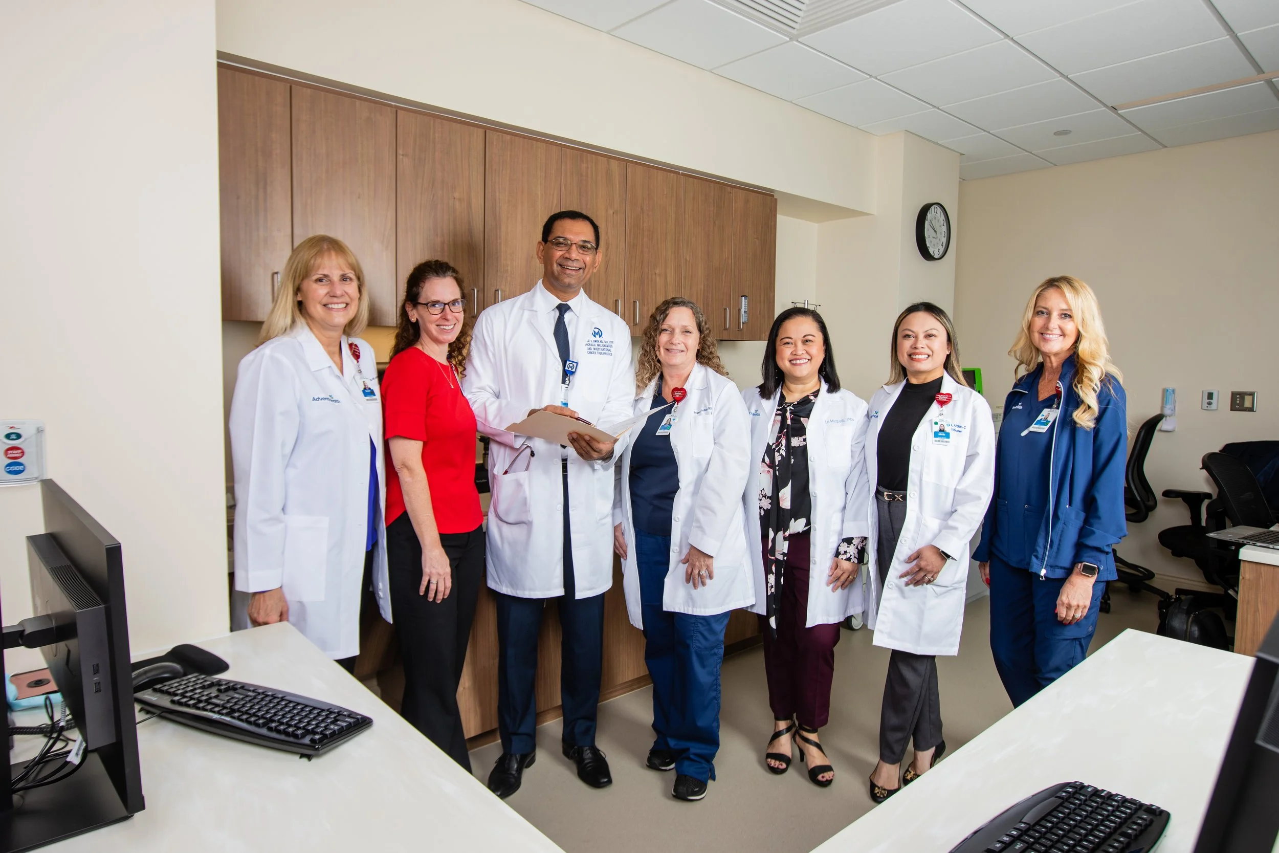 Group of seven diverse medical professionals, including doctors and nurses, standing together in a hospital or medical office, smiling for the camera.