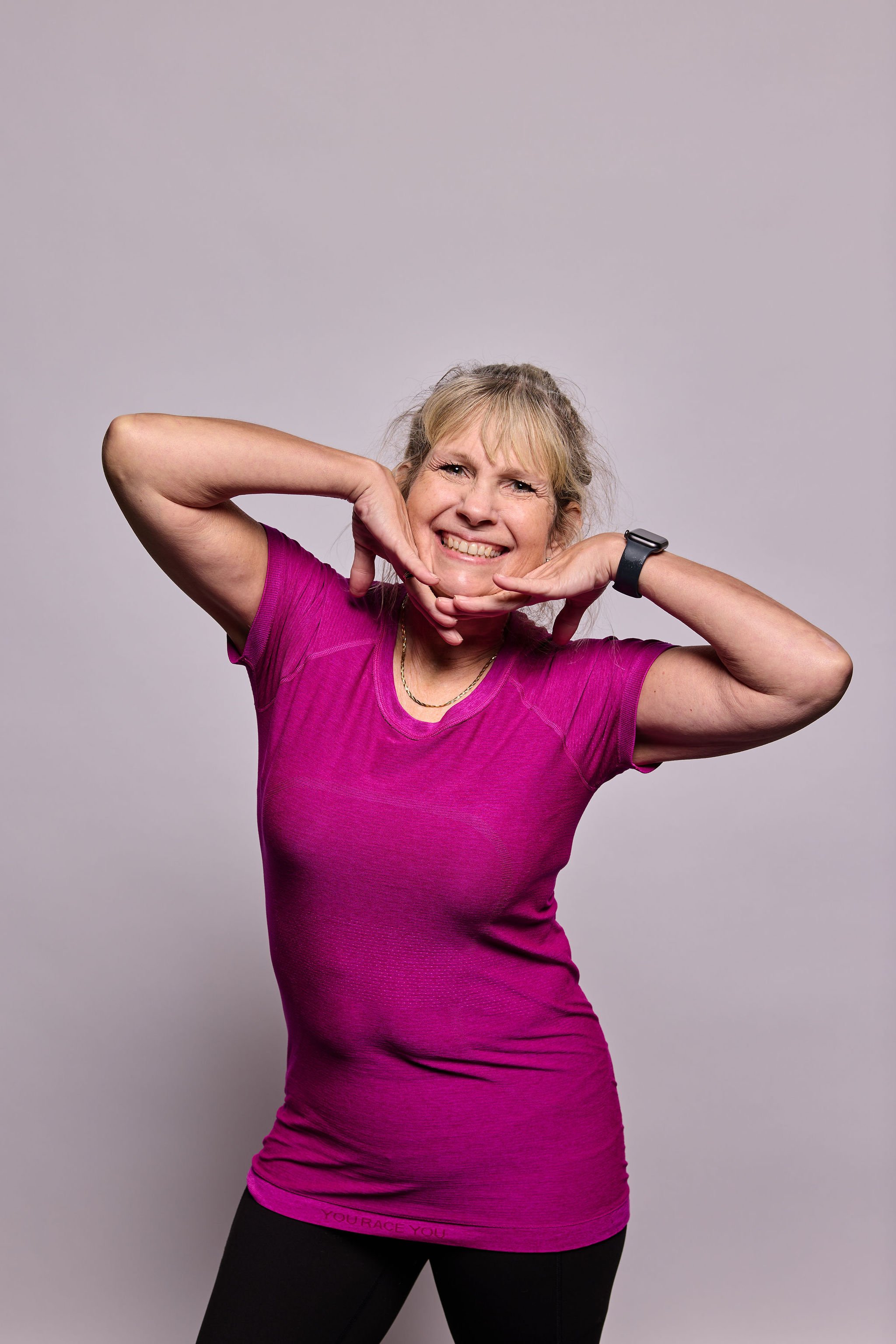 A woman with blonde hair in a ponytail, wearing a pink athletic shirt and black pants, smiling with her teeth showing while posing with her hands framing her face against a gray background.