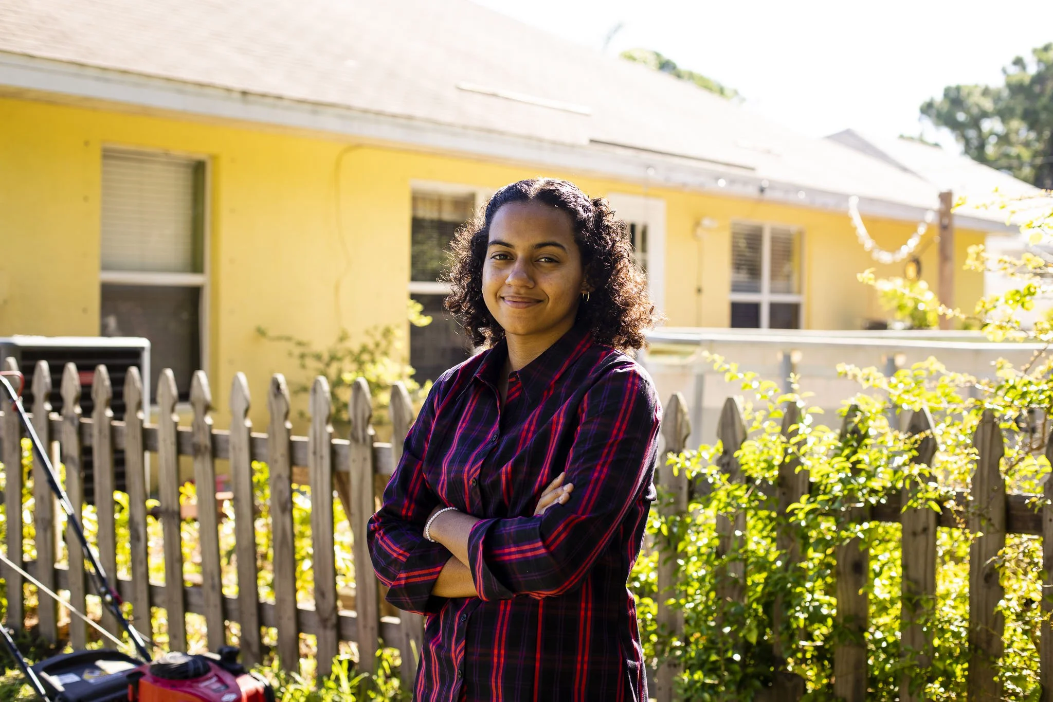 Young woman with curly hair standing outside in sunlight, with arms crossed, in front of a wooden fence and yellow house, smiling.