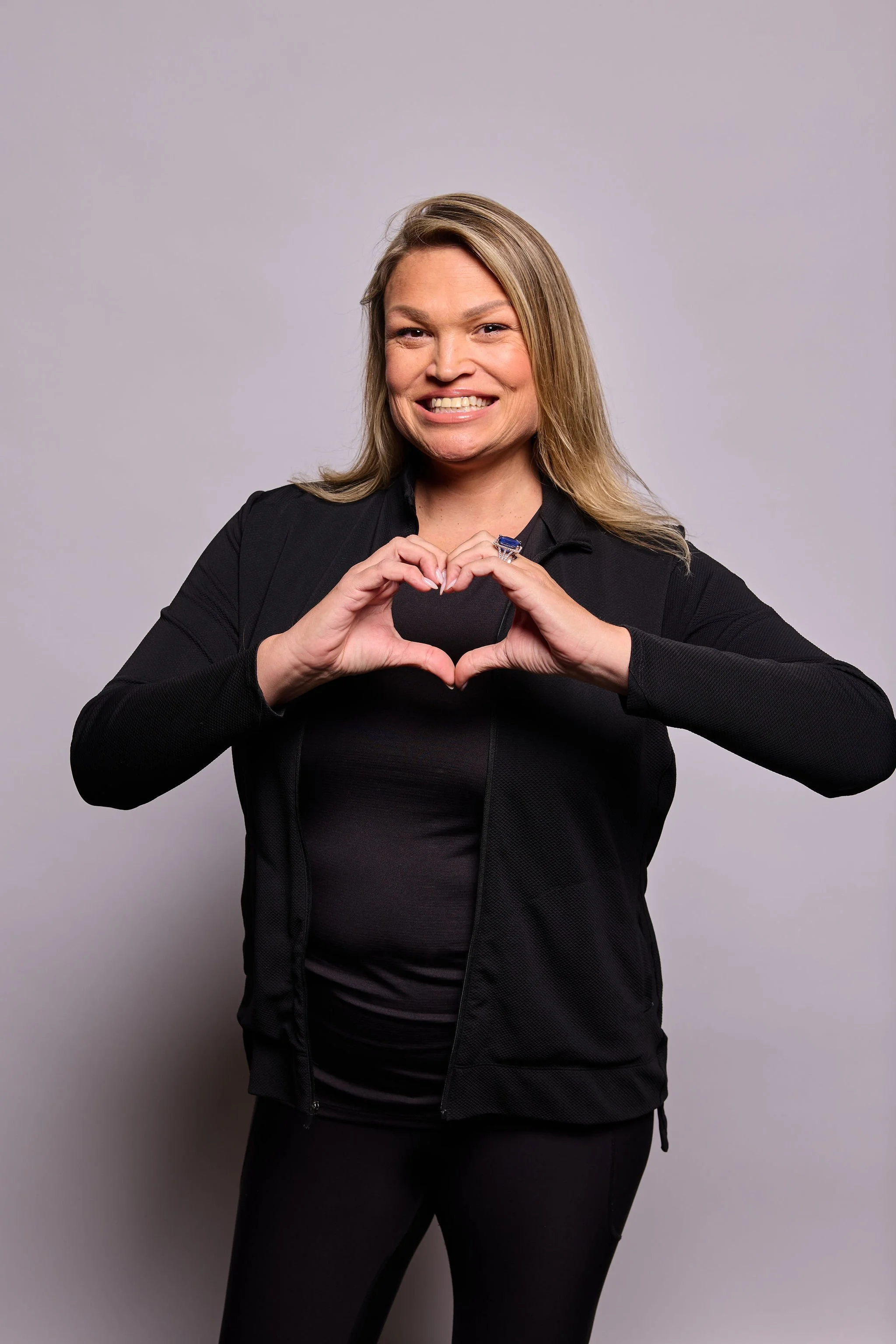 Woman in black athletic clothing making a heart shape with her hands, smiling, standing against a plain gray background.